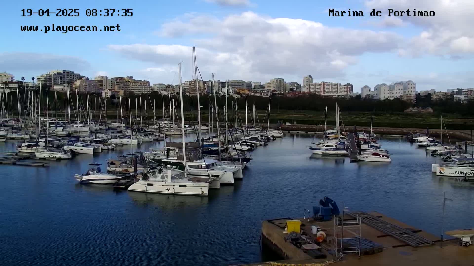 A marina filled with numerous sailboats and motorboats is situated in front of a city skyline under a partly cloudy sky.