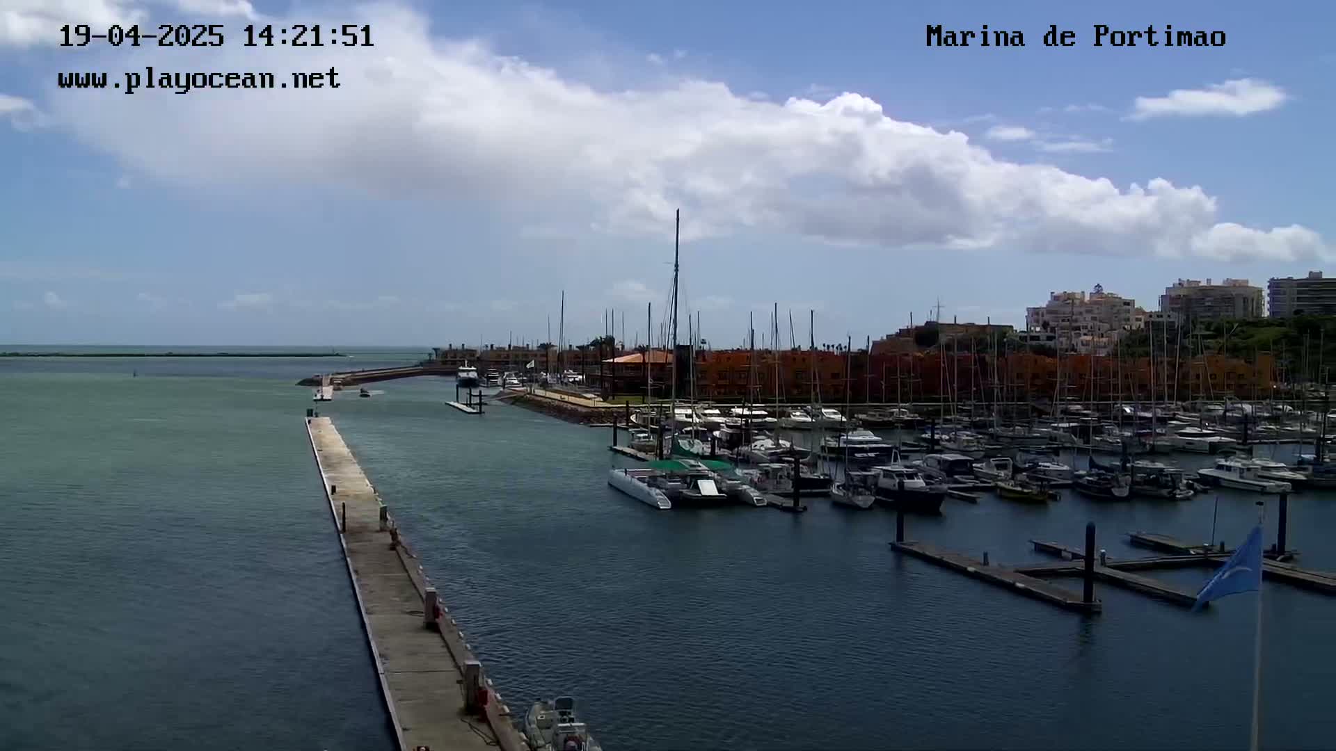 A marina filled with numerous boats is seen on a partly cloudy day, with a long pier extending into the calm water.