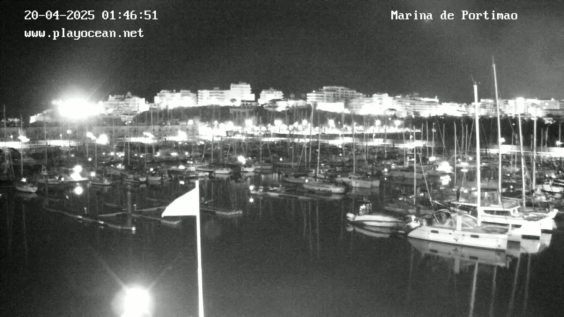 A nighttime, low-light view of a marina filled with numerous boats and yachts, backed by a shoreline of illuminated buildings.