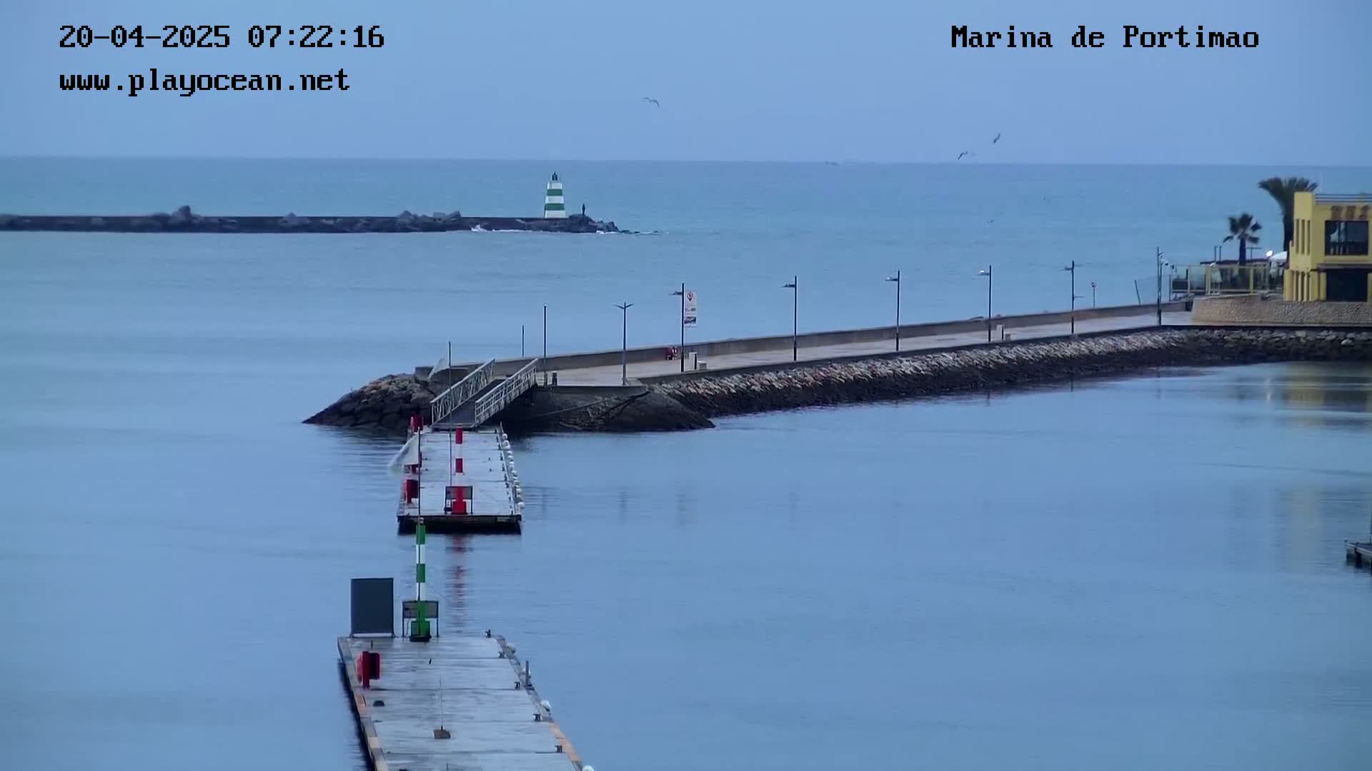 A calm, overcast day shows a marina with a long walkway and docks extending into the water, a lighthouse visible in the distance.