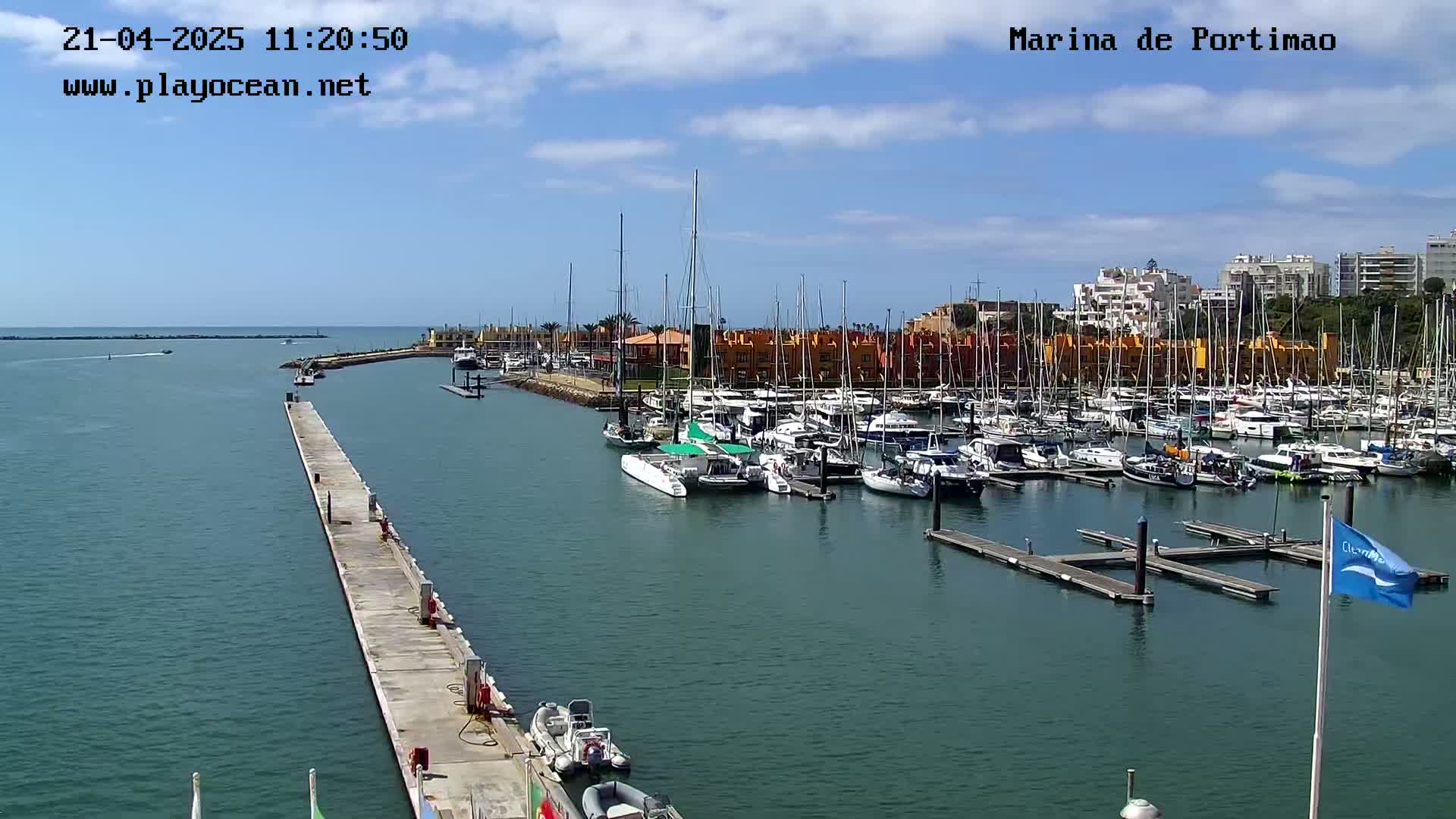 A marina filled with numerous sailboats and motorboats is seen on a sunny day, with calm waters and a long pier extending into the harbor.