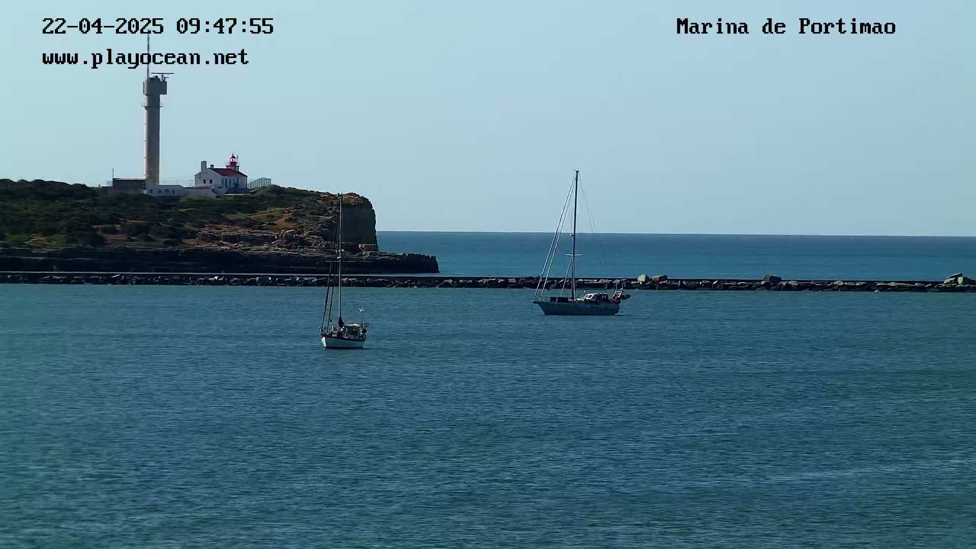 Two sailboats are anchored in calm waters near a rocky coast featuring a lighthouse and a tall tower under a clear sky.