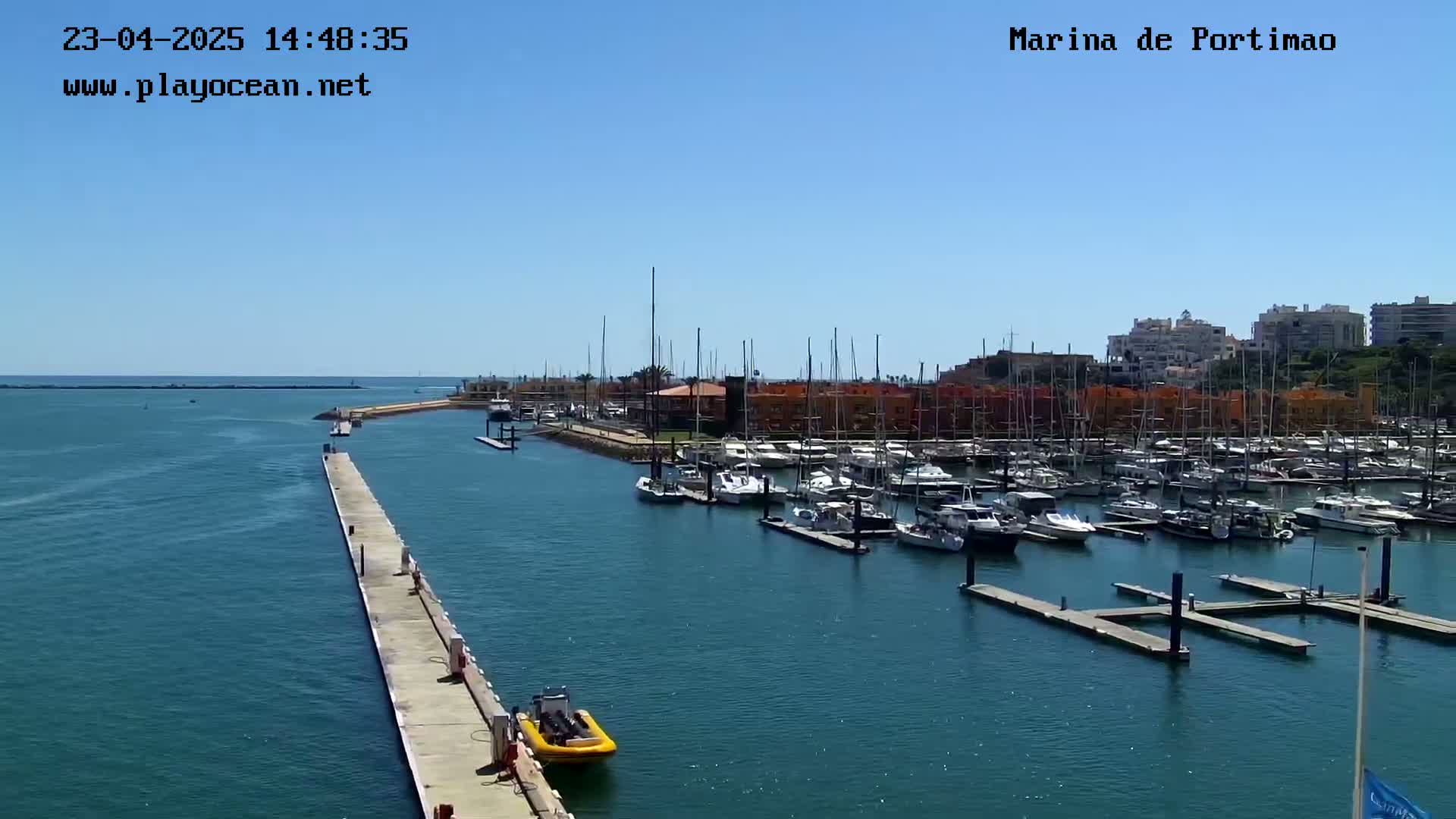 A marina filled with numerous boats and yachts is seen on a sunny day, with a long pier extending into the calm water.