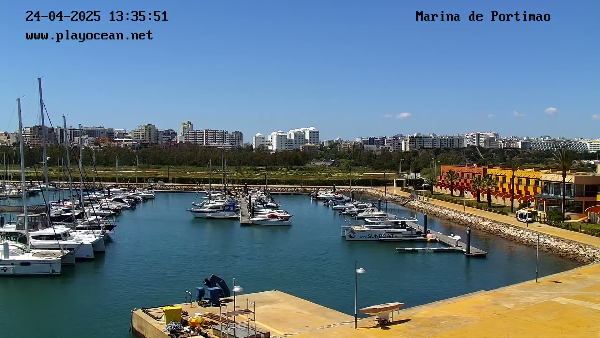 A marina filled with sailboats and motorboats is situated near a waterfront building under a clear blue sky.