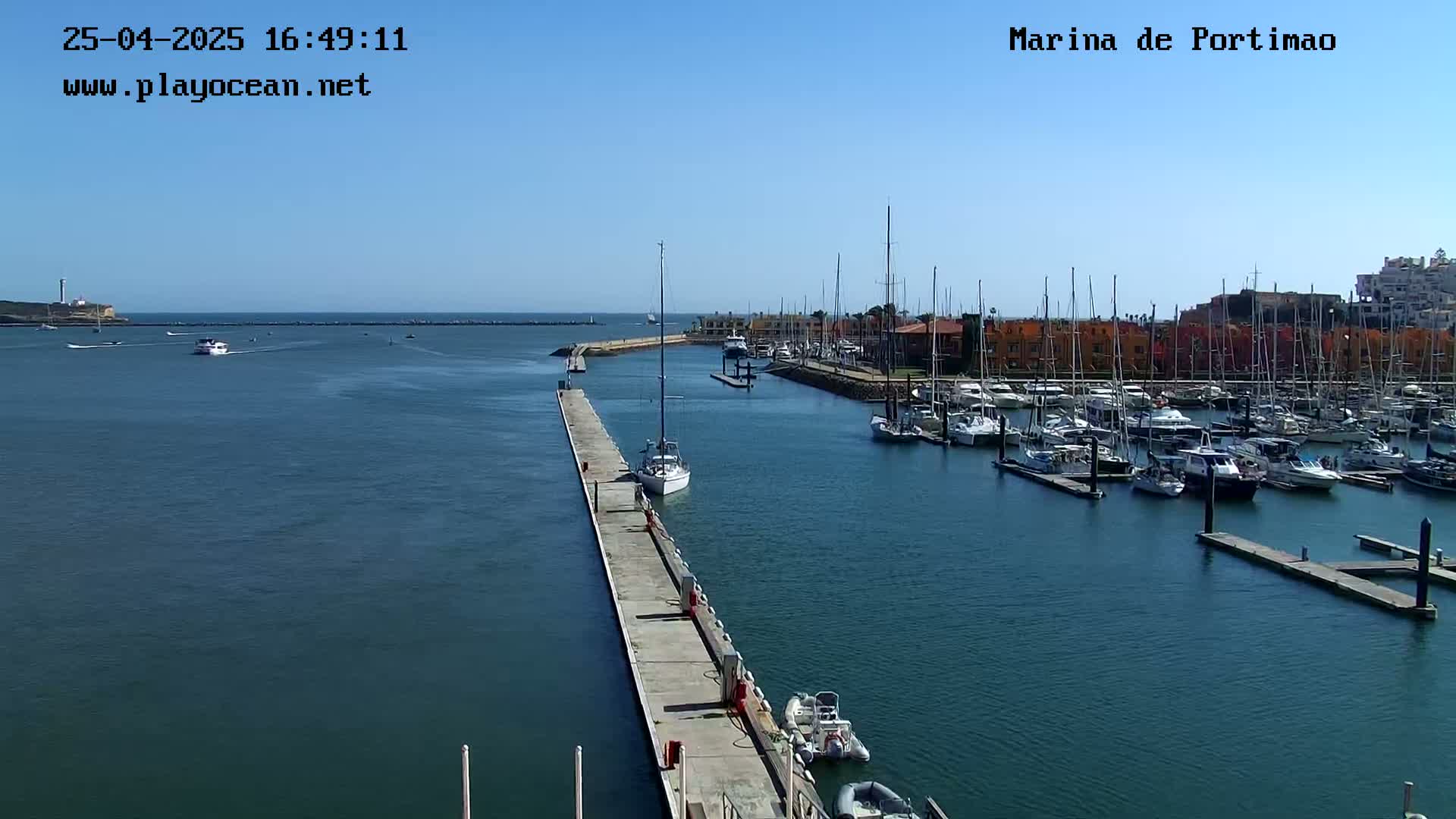 A marina filled with numerous sailboats and motorboats is seen on a sunny day, with a long pier extending from the shore into the calm water.