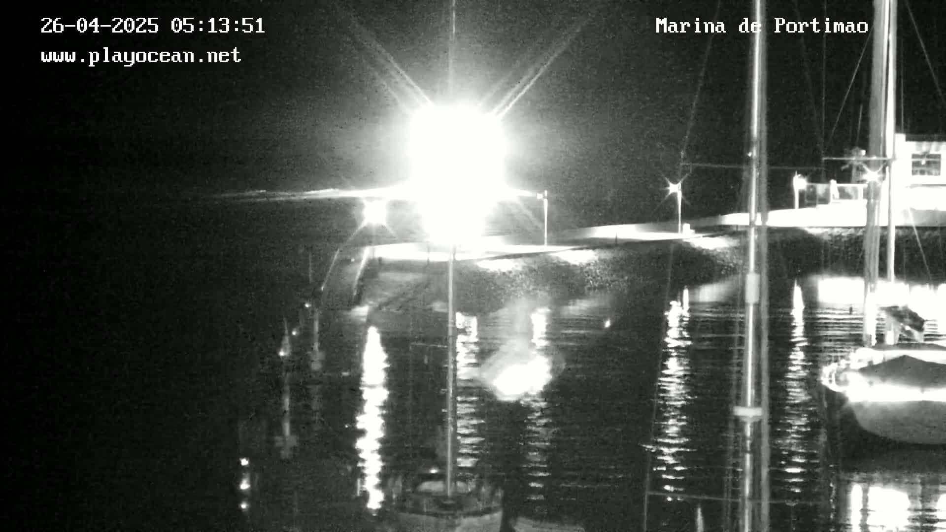 A nighttime view of a marina with boats moored, illuminated by bright lights reflecting on calm water.