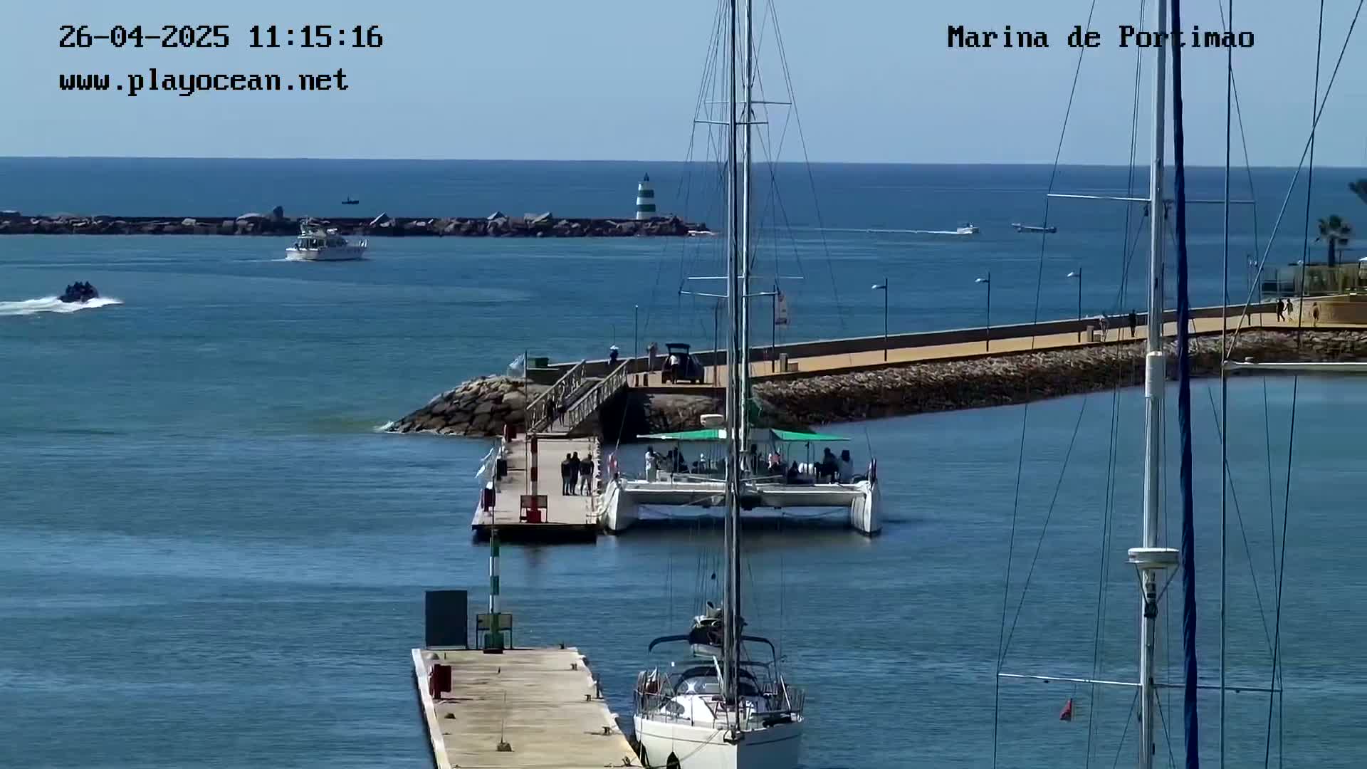 A marina scene on a sunny day shows several boats, including a catamaran at a dock with people on board, and other sailboats in the water.