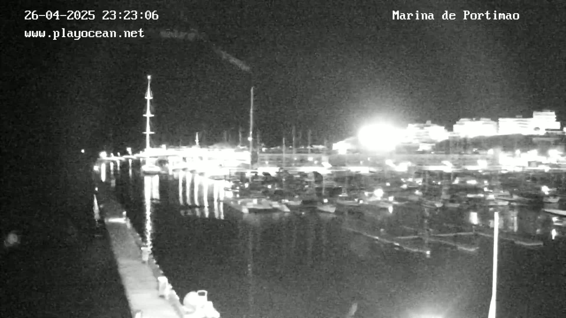 A nighttime, black-and-white view of a marina filled with boats, with a calm body of water reflecting the lights from the boats and surrounding buildings.