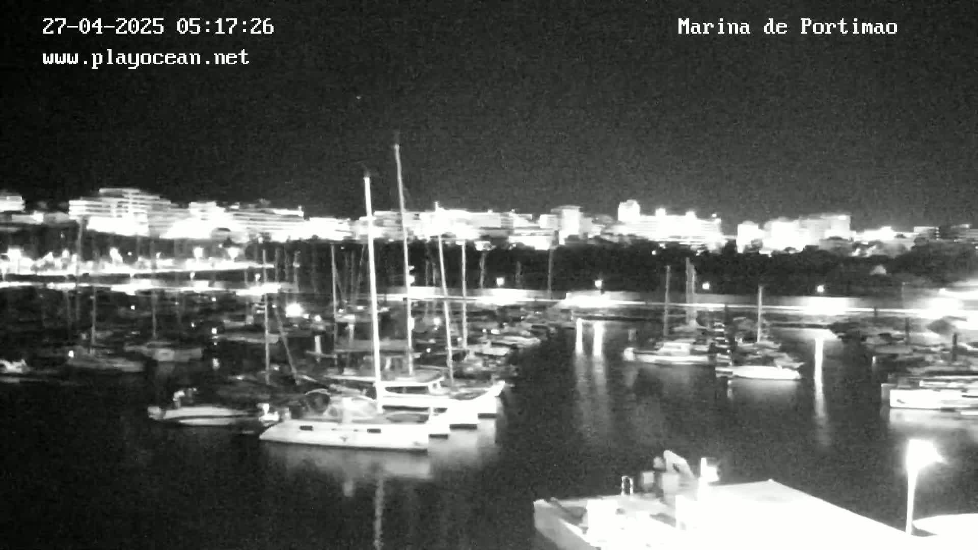 A nighttime view of a marina filled with boats, with a brightly lit city skyline in the background.