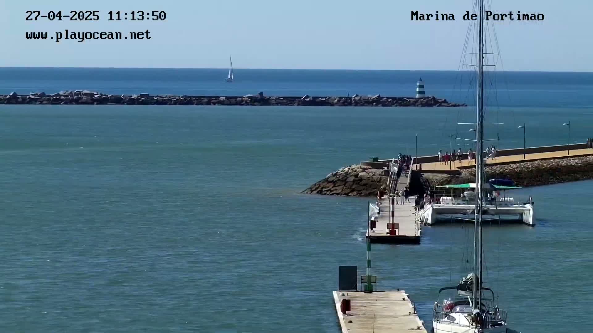 A marina with calm, blue water, a sailboat at dock, and a catamaran, under sunny skies.