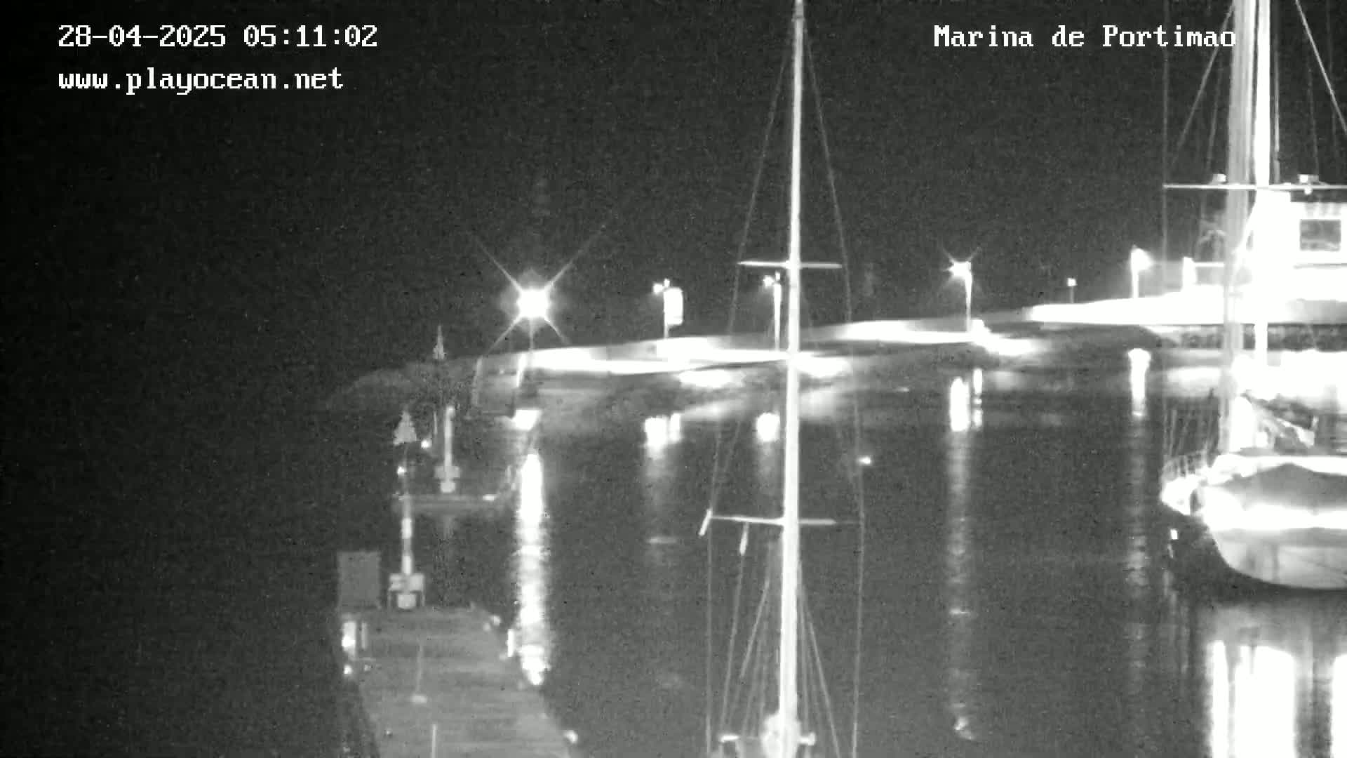 A nighttime view of a marina with several boats docked, illuminated by lights reflecting on the calm water.