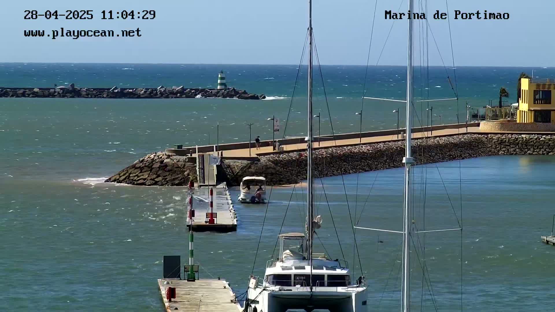 A marina with a catamaran in the foreground, a small motorboat leaving a dock, and a calm, sunny day with a slight wind visible on the water.
