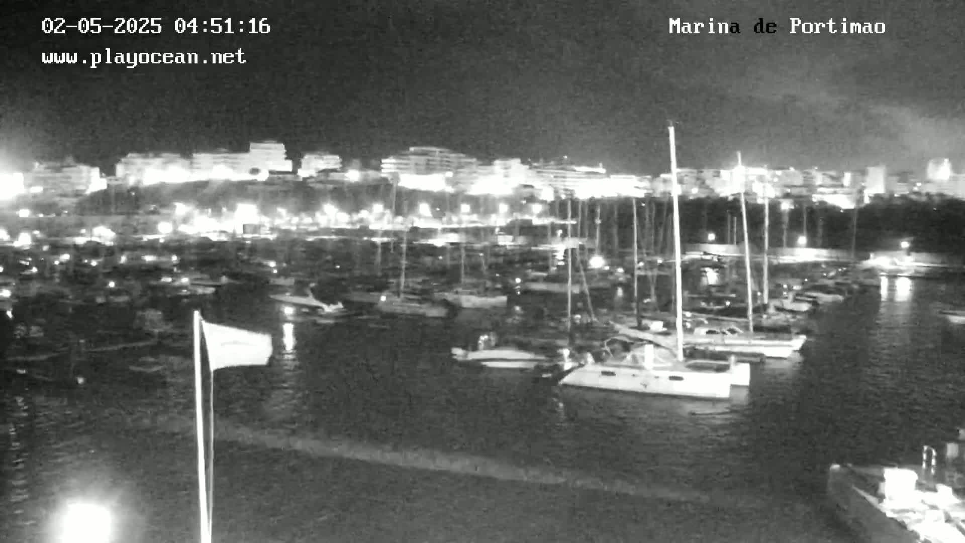 A nighttime, low-light view of a marina filled with docked sailboats, with a brightly lit town visible in the background.