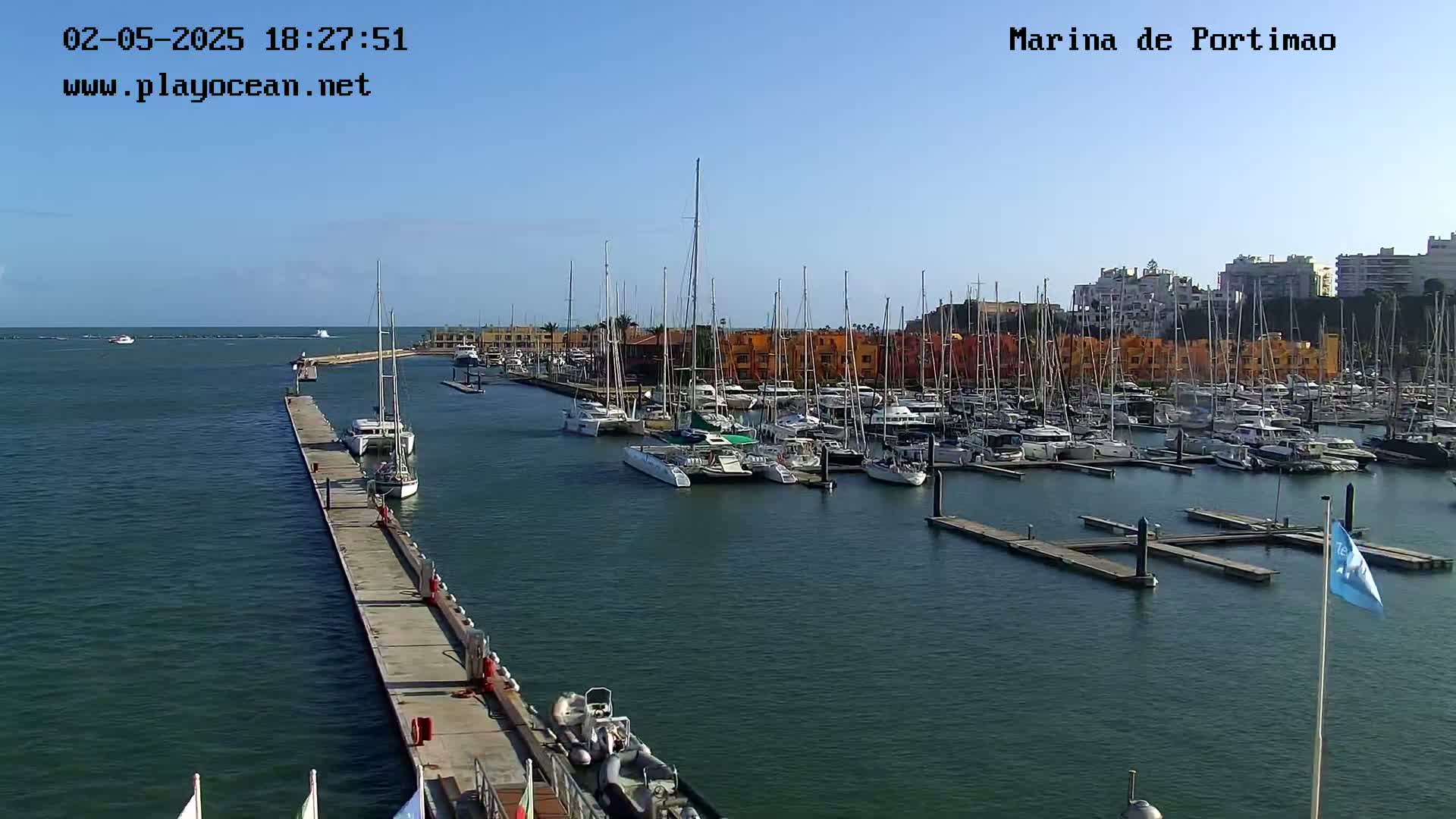 A marina filled with numerous sailboats and yachts is shown on a sunny day, with a long concrete pier extending into the calm water.