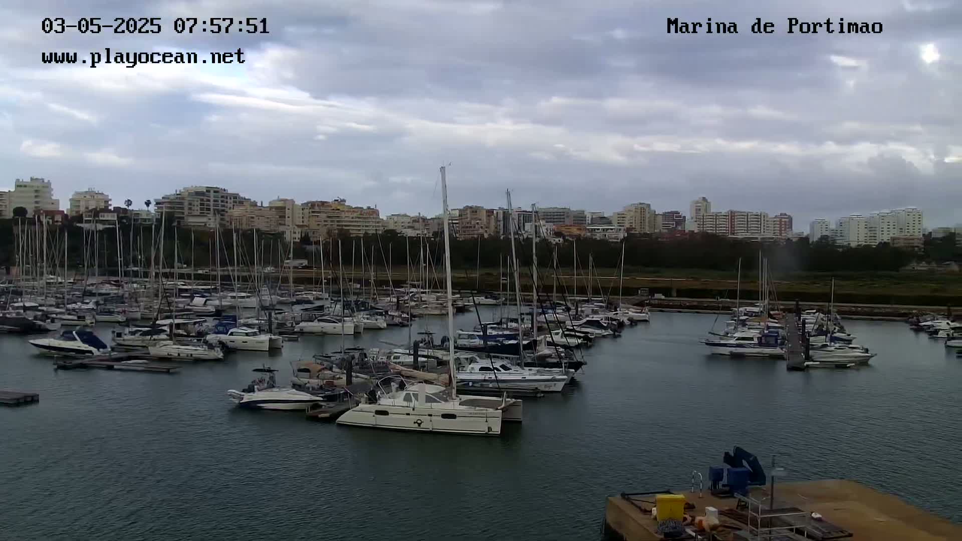 A marina filled with numerous sailboats and motorboats is situated under a cloudy sky, with a cityscape visible in the background.