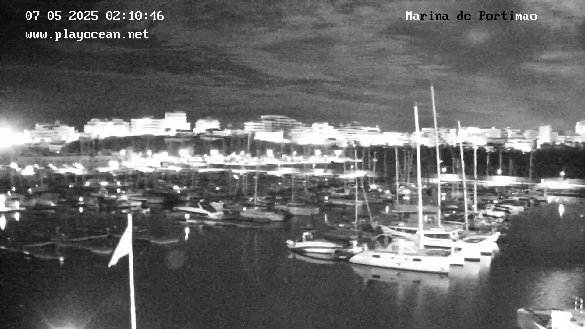 A nighttime, low-light view of a marina filled with numerous boats and yachts, with a city skyline illuminated in the background under a cloudy sky.