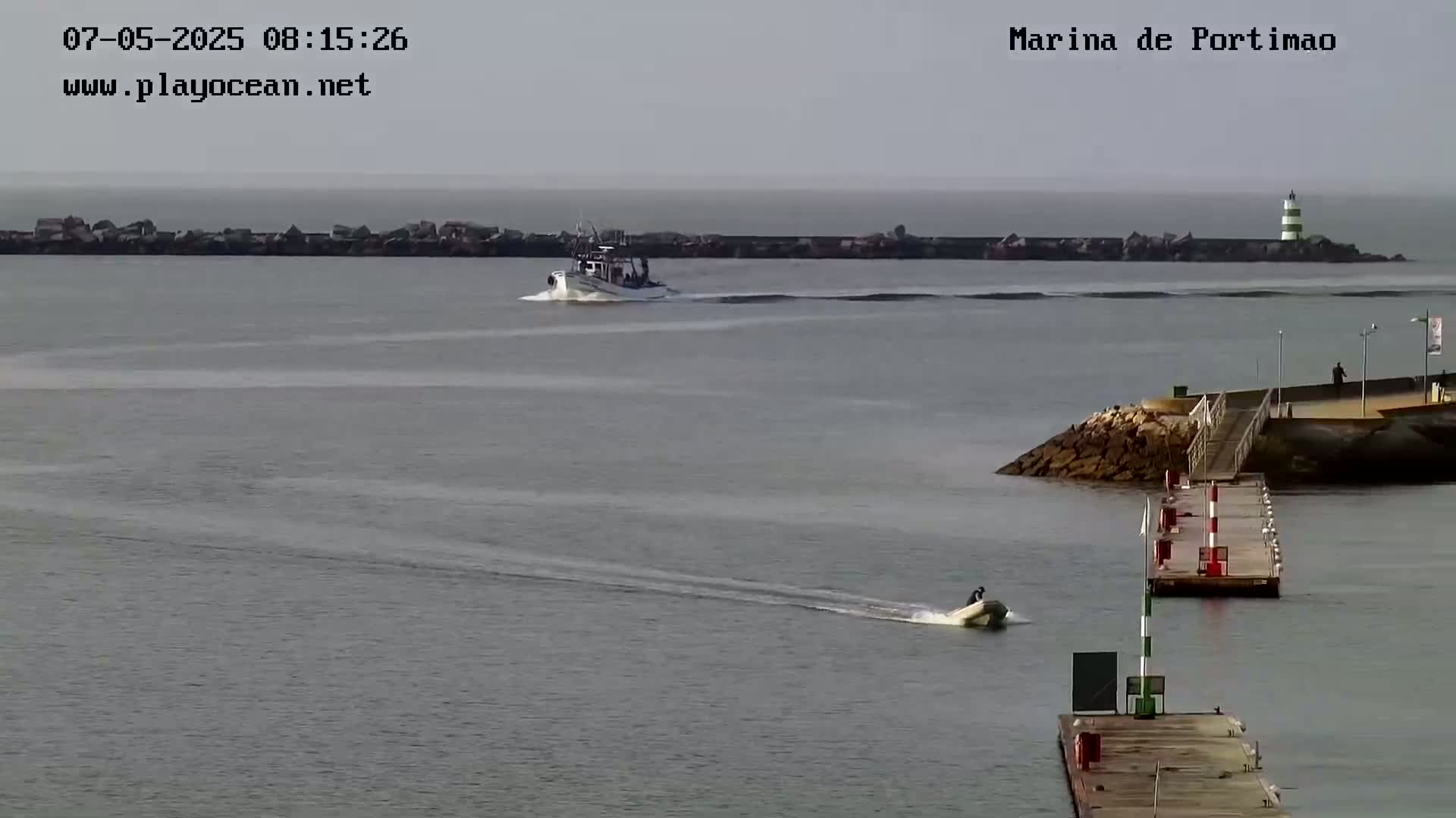 A calm, gray day shows a small motorboat speeding across the water toward a pier, while a larger boat passes in the distance near a breakwater with a lighthouse.