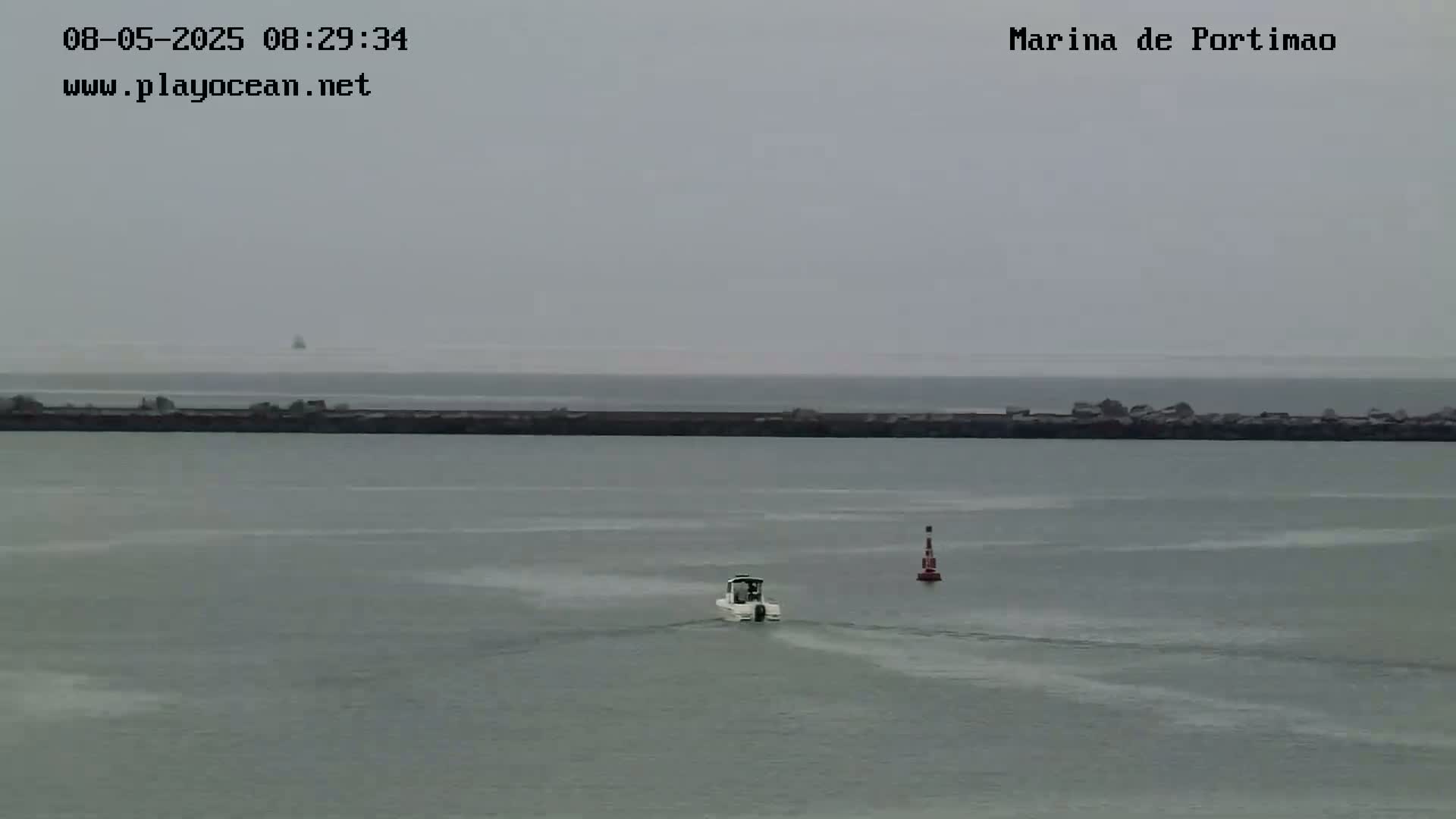 A small motorboat travels across a calm, gray body of water past a breakwater on an overcast day.