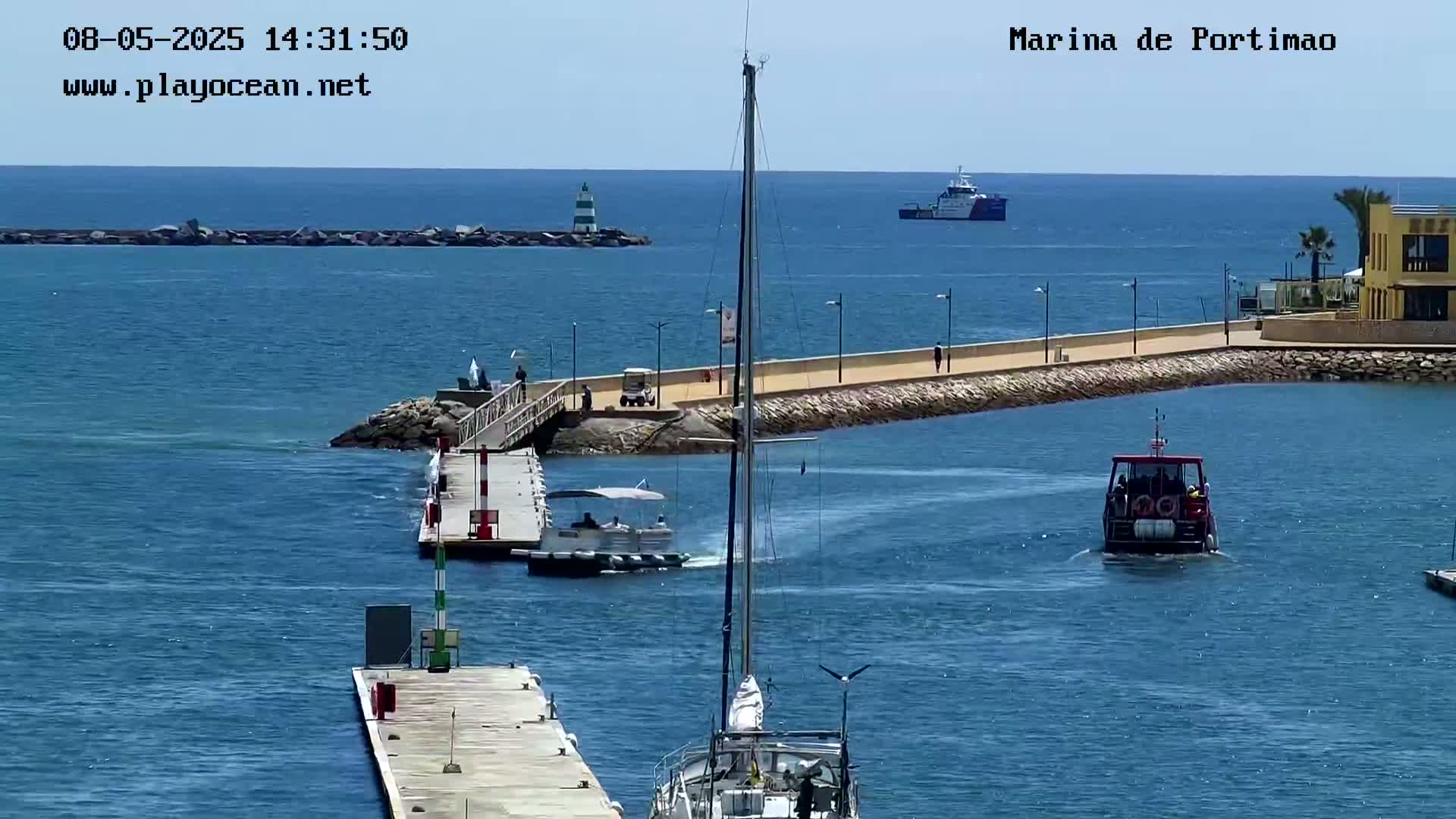 A marina with calm, blue water shows several boats, including a sailboat in the foreground, a small motorboat near a pier, and a larger vessel further out in the harbor on a sunny day.