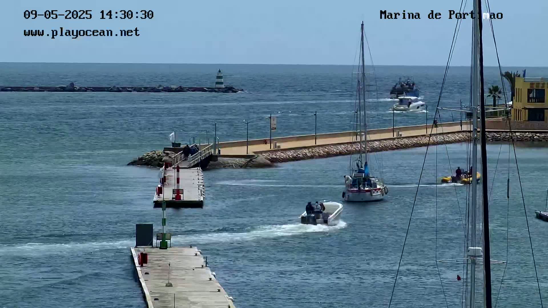 A marina scene on a sunny day shows several boats in the water, with a small powerboat passing near a dock and larger sailboats moored nearby.