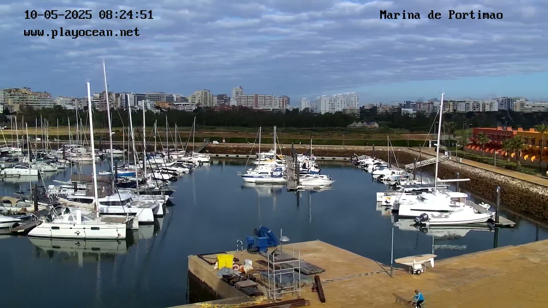 A marina filled with numerous sailboats and motorboats is situated near a town under a partly cloudy sky.