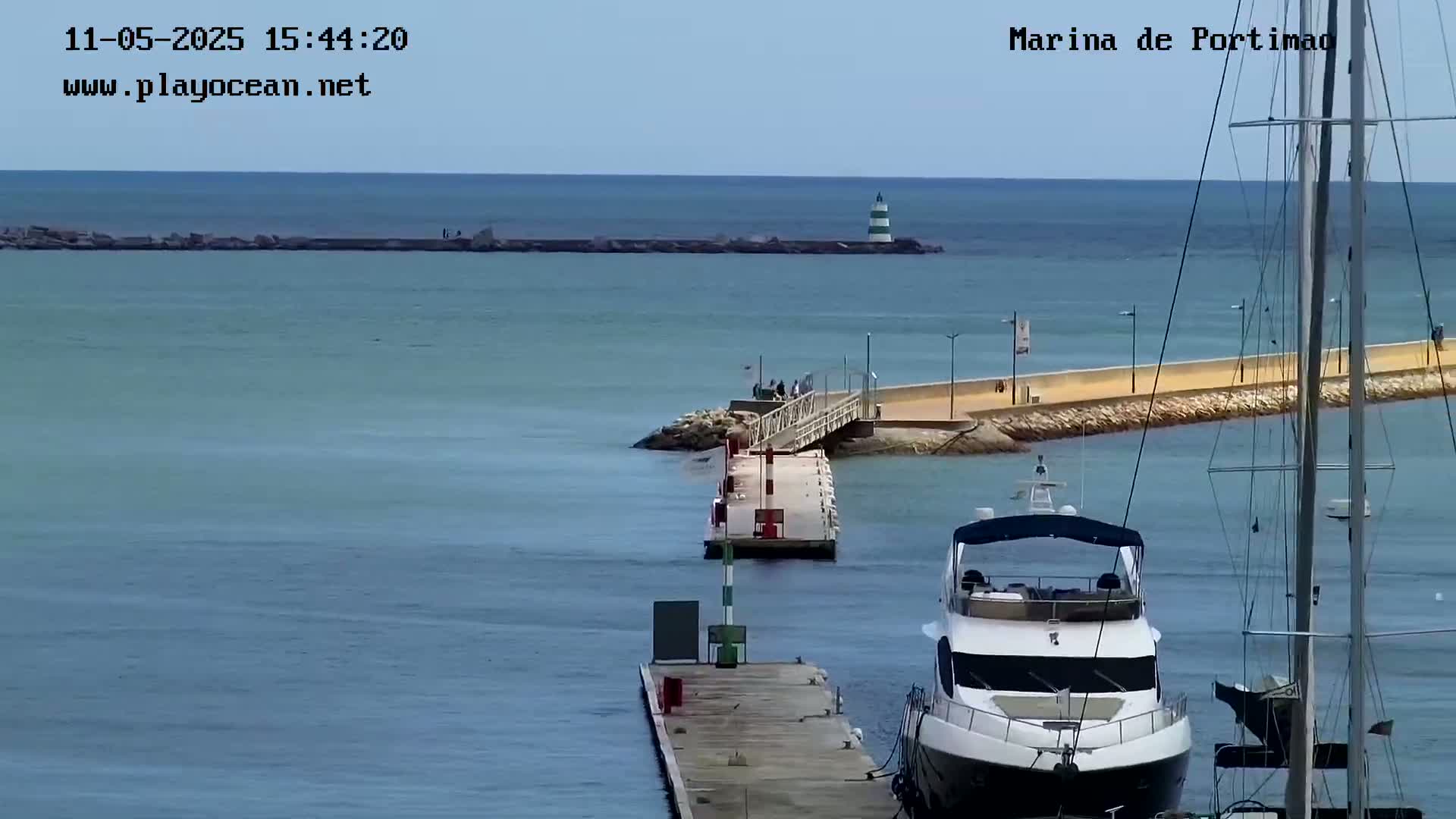 A yacht is docked near a long pier extending into calm, blue water, with a lighthouse visible in the distance under a clear sky.