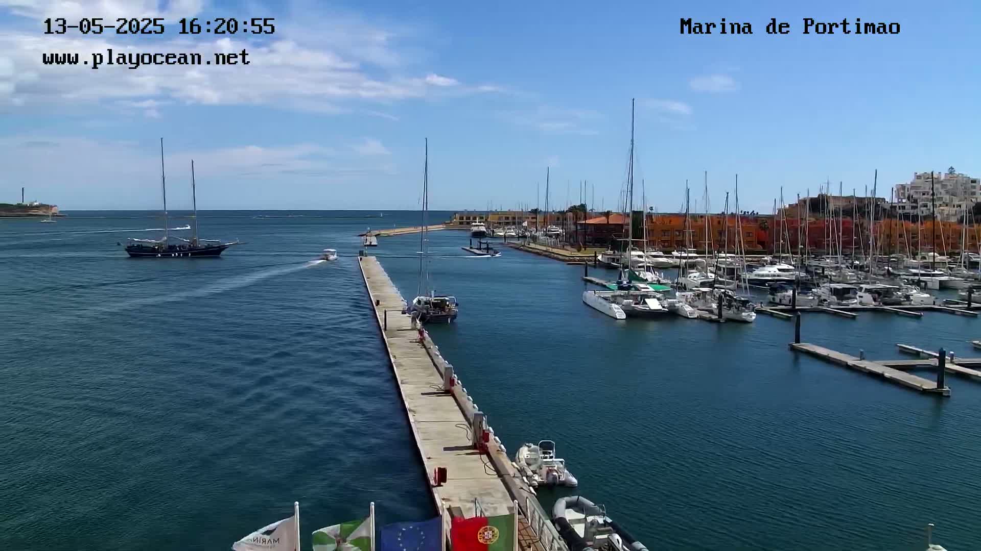 A marina filled with numerous sailboats and yachts is shown on a sunny day, with a large sailing ship visible in the distance.