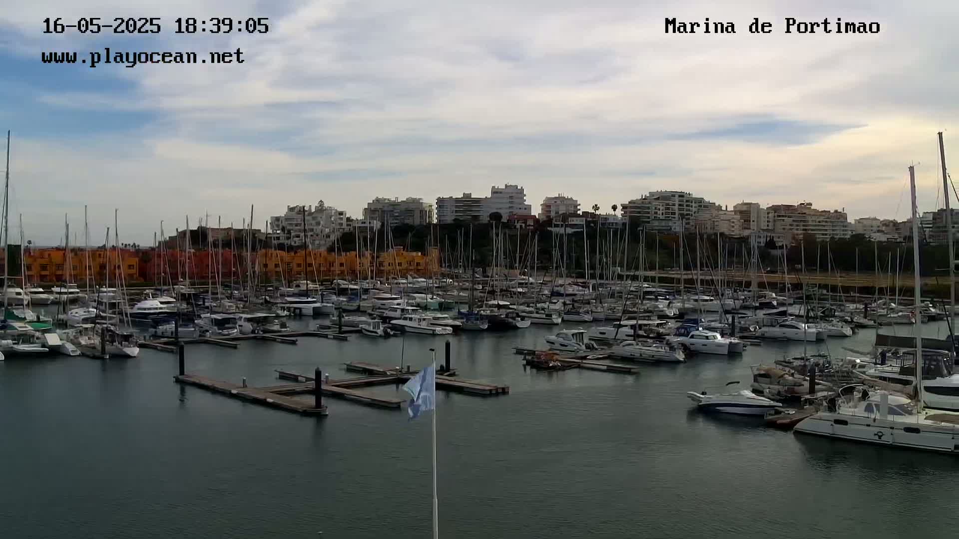 A marina filled with many boats is situated in front of a line of buildings under a partly cloudy sky.