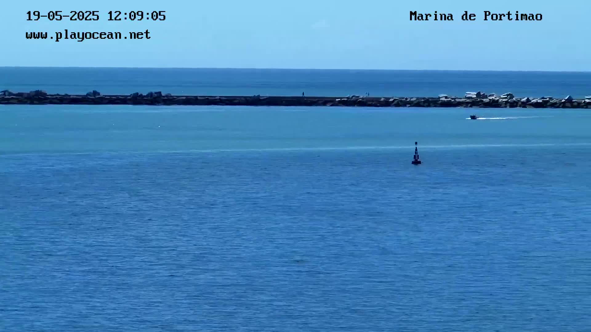 A small boat is visible on calm, blue water beyond a long, dark breakwater.
