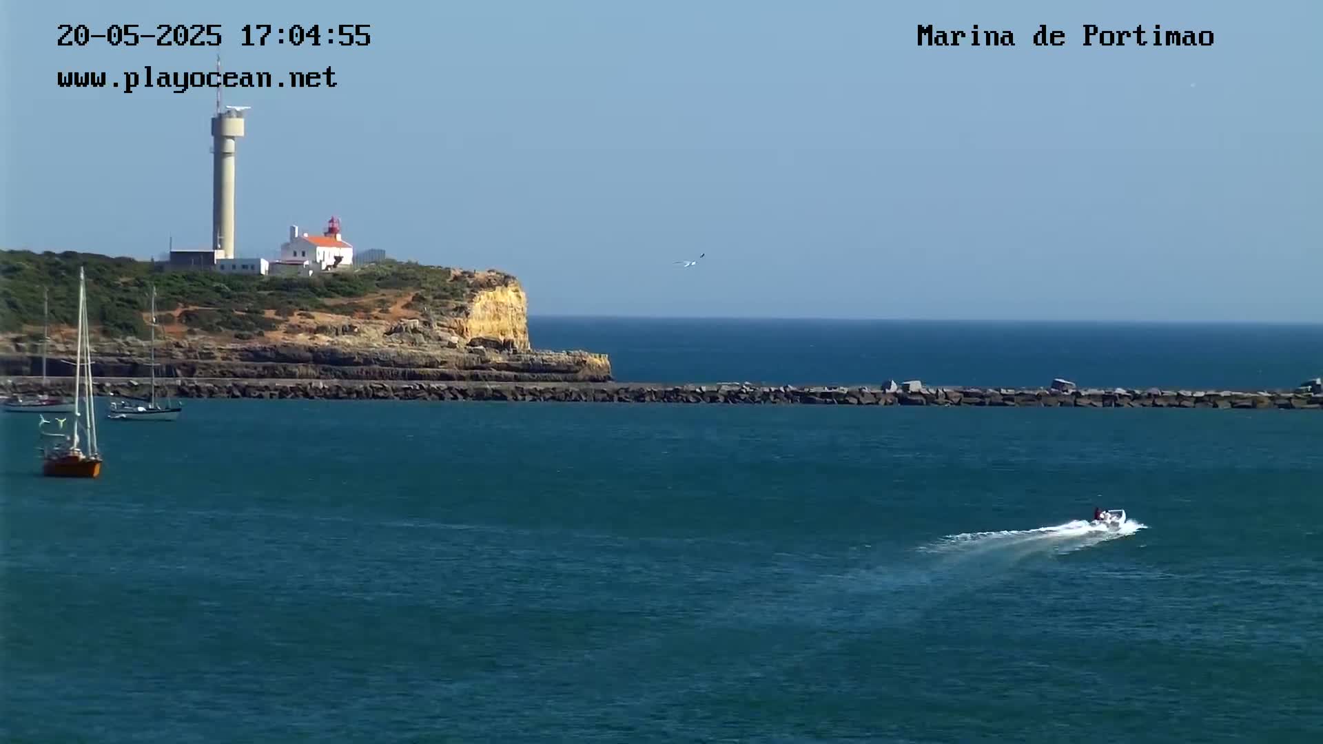 A coastal scene shows a lighthouse on a cliff overlooking a calm body of water with several sailboats and a motorboat, under a clear sky.