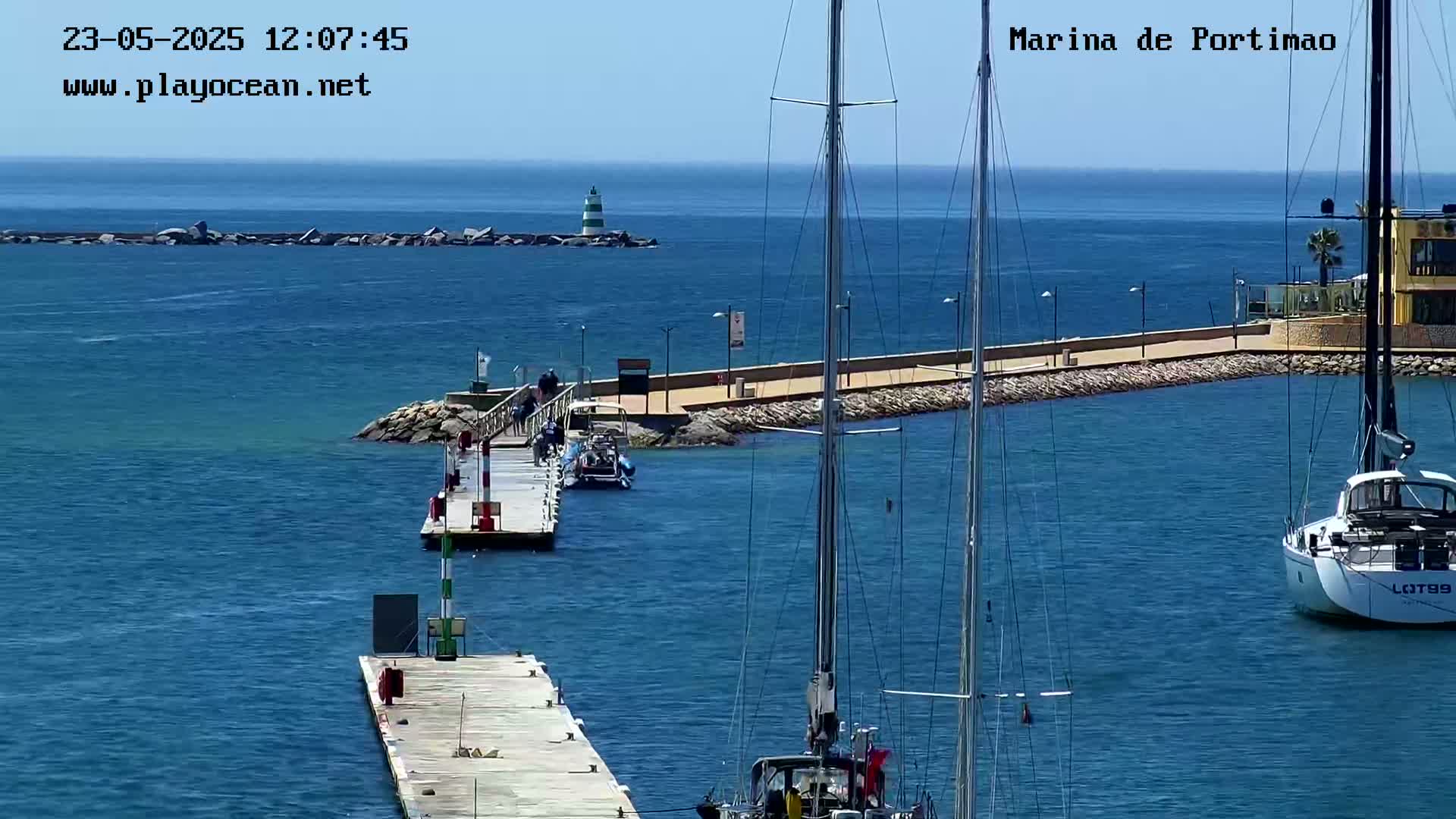 A marina with calm blue water, several sailboats, a small motorboat docked at a pier, and a lighthouse in the distance under sunny skies.