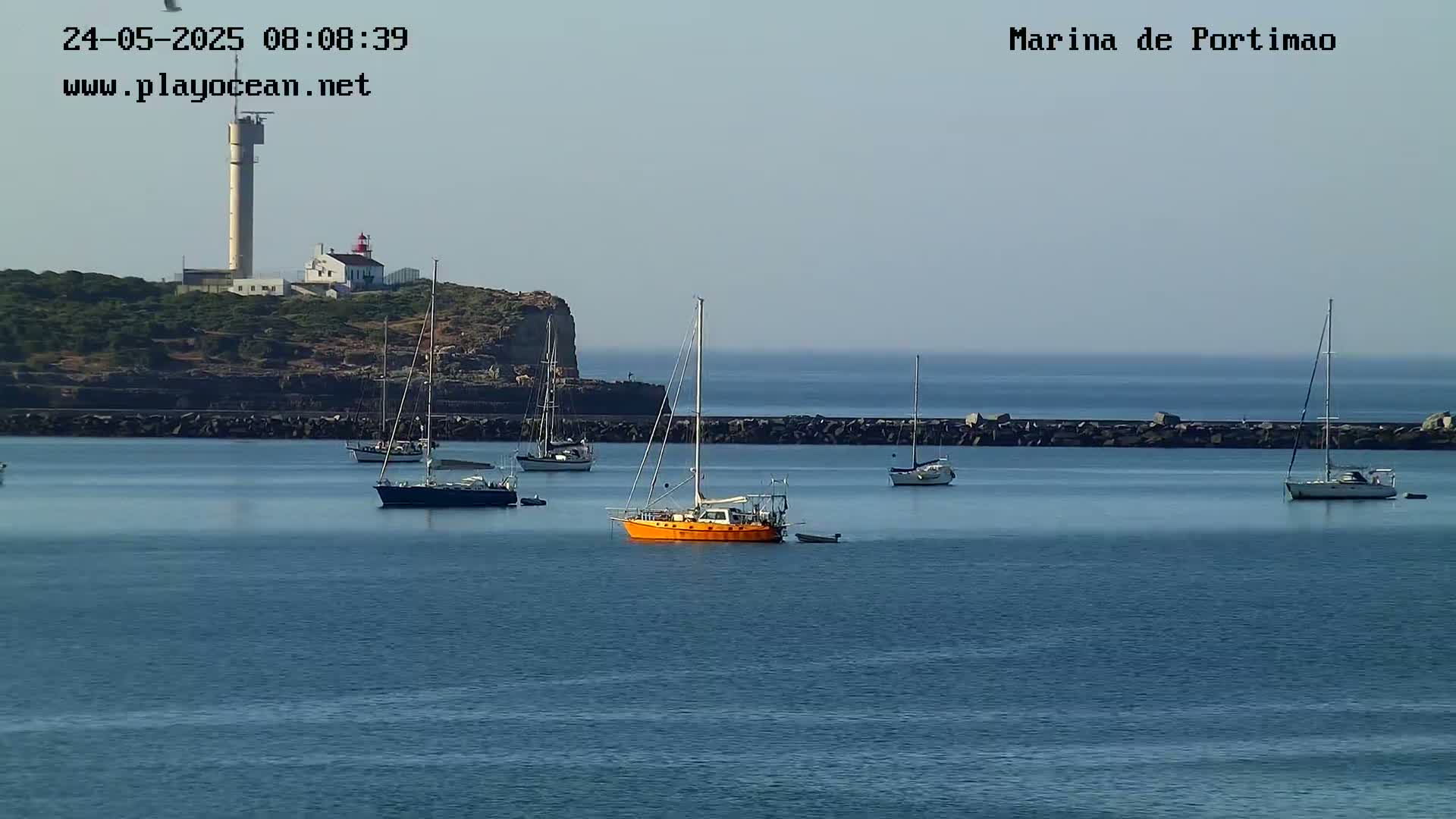 Several sailboats are moored in a calm harbor next to a rocky headland with a lighthouse under a clear sky.