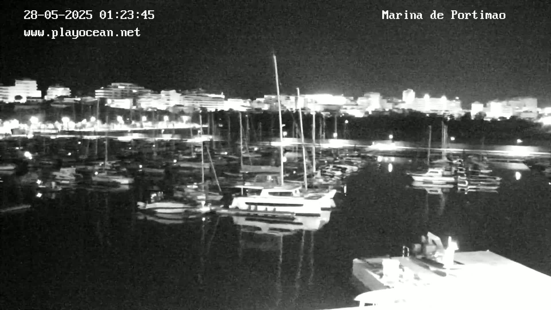 A nighttime view of a marina filled with boats, reflecting the lights of a city in the calm water.