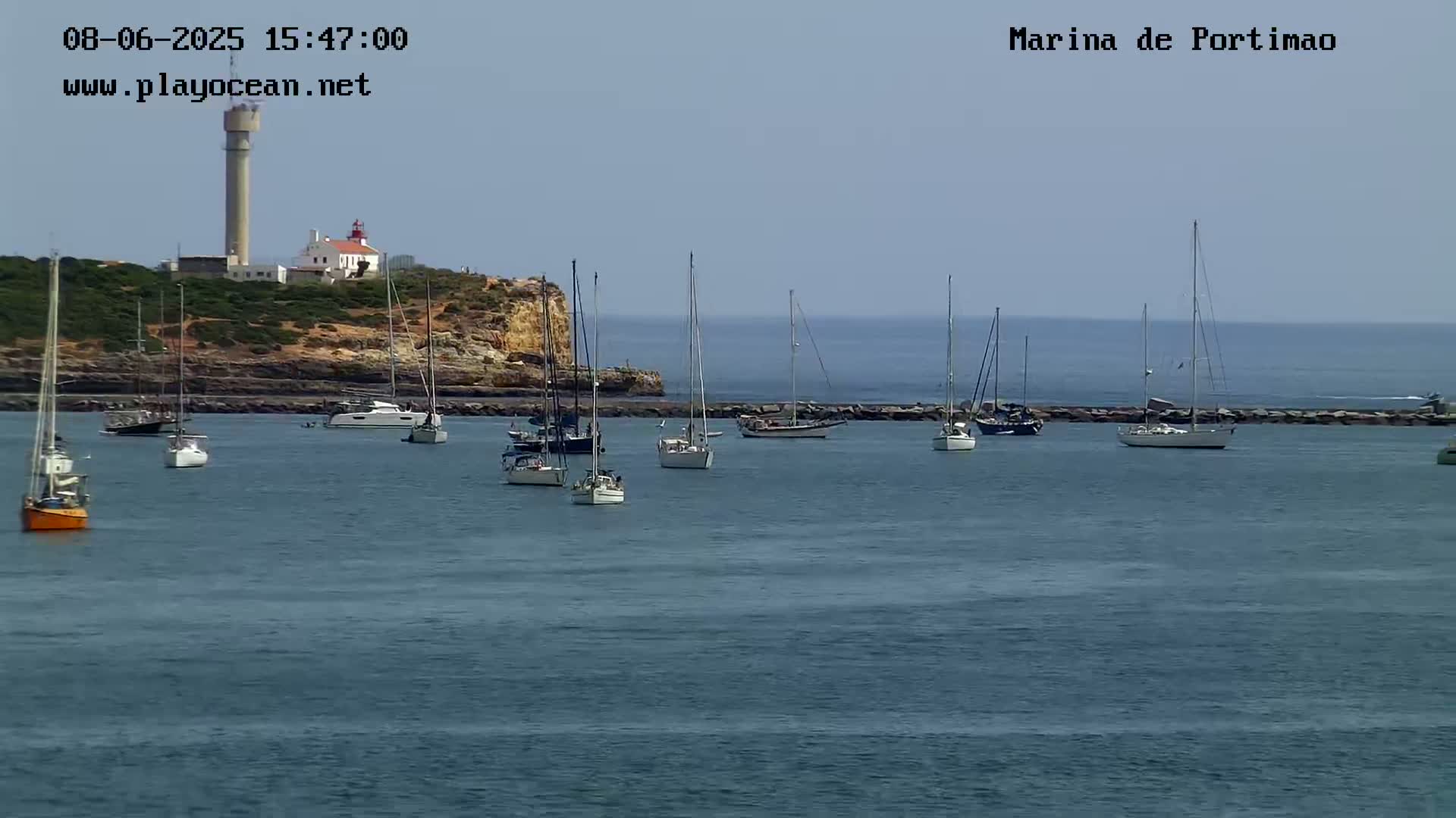 Several sailboats are moored in a calm harbor next to a rocky coastline featuring a lighthouse and a tall tower under a clear sky.