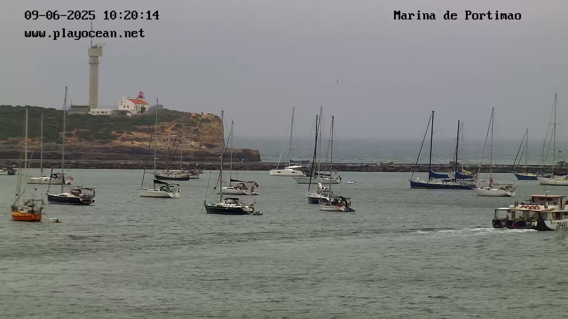 Numerous sailboats and a small passenger boat are moored in a calm harbor, with a lighthouse and tower visible on a nearby cliff under an overcast sky.
