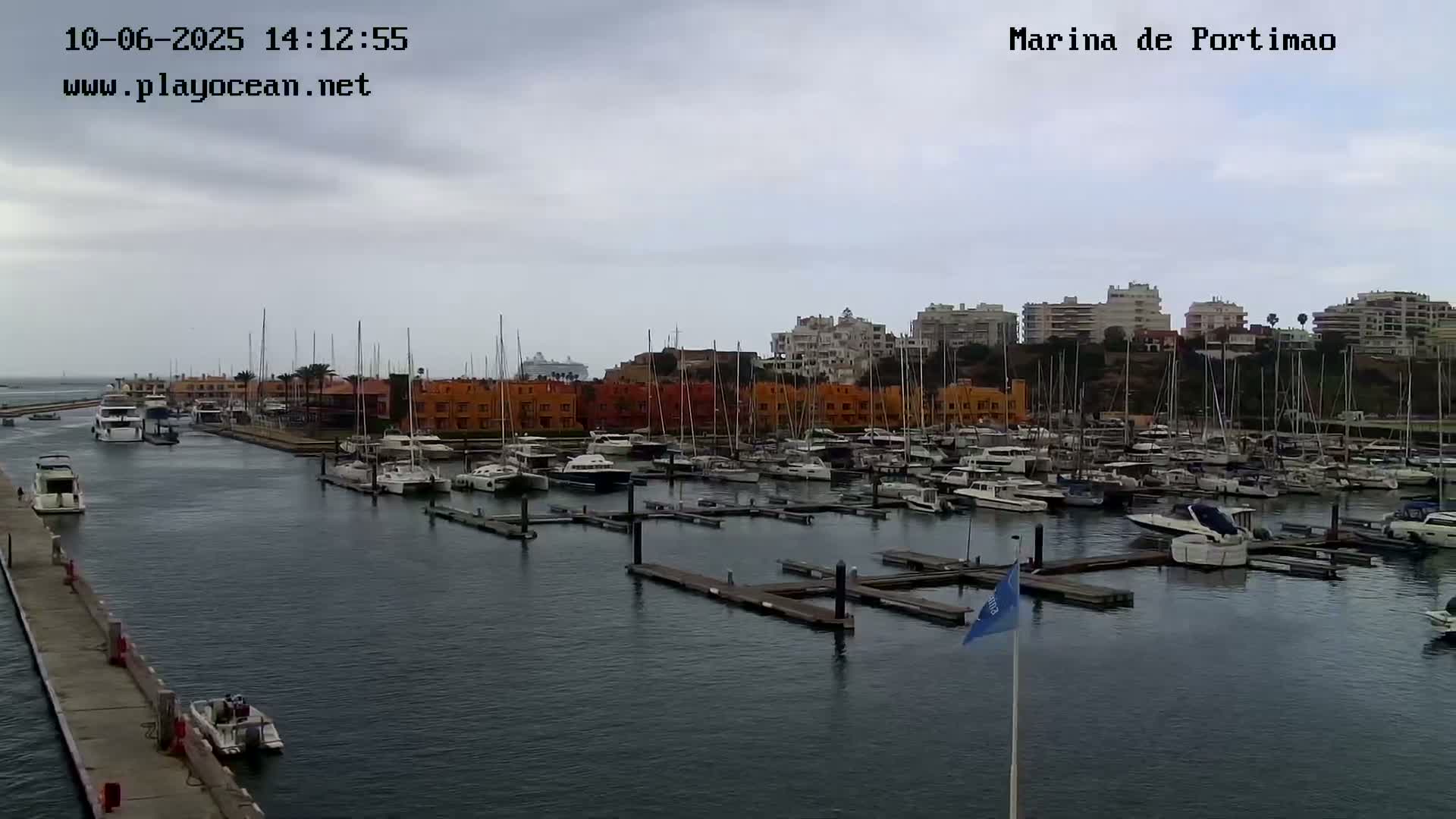 A marina filled with numerous boats and yachts is situated under an overcast sky, with buildings lining the shoreline in the background.