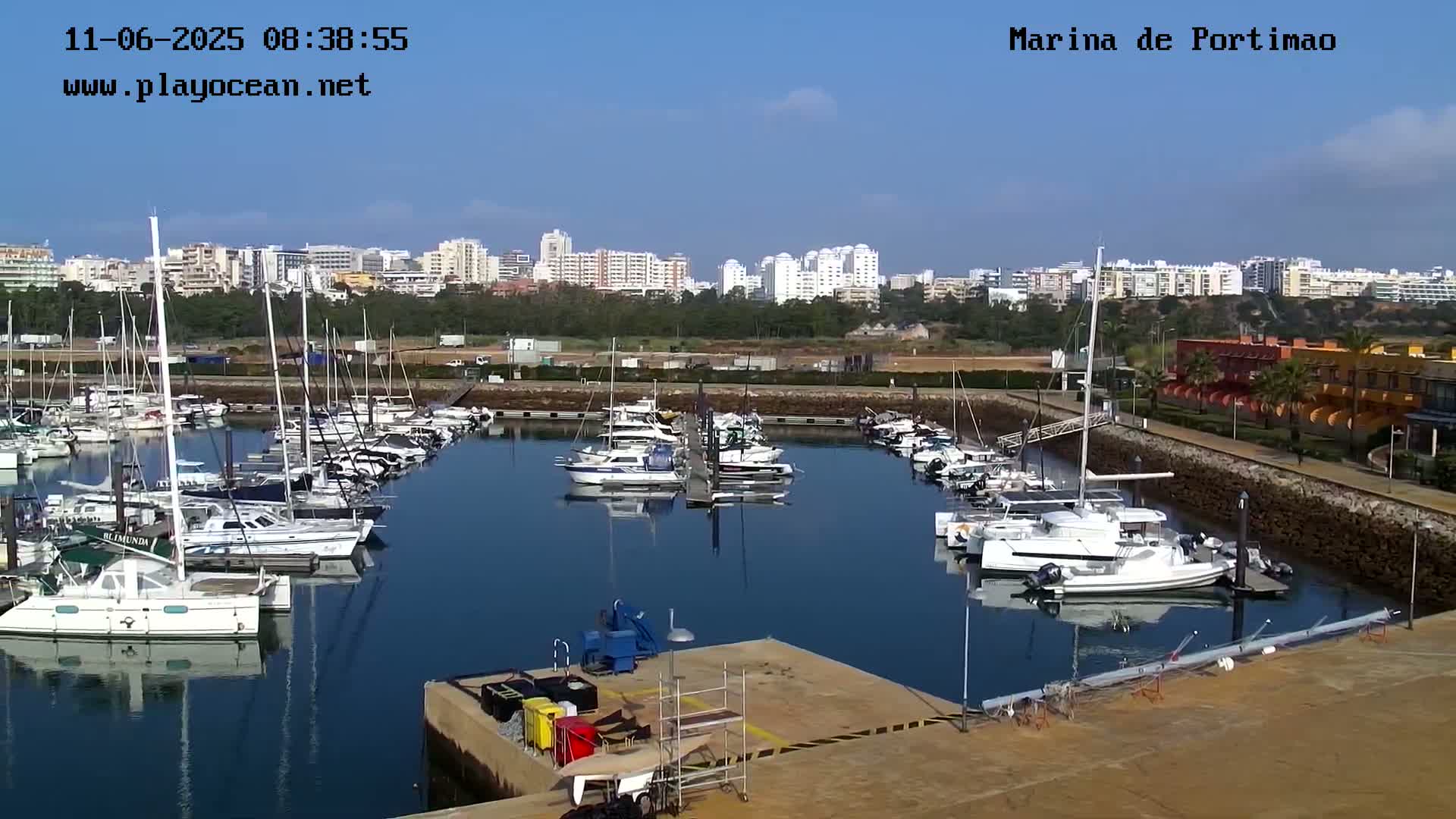 A marina filled with numerous boats and yachts is situated in front of a coastal city under a clear blue sky.
