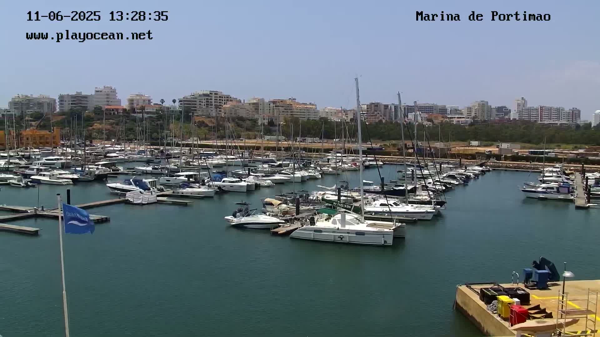A marina filled with numerous boats and yachts is situated near a city under a clear, sunny sky.