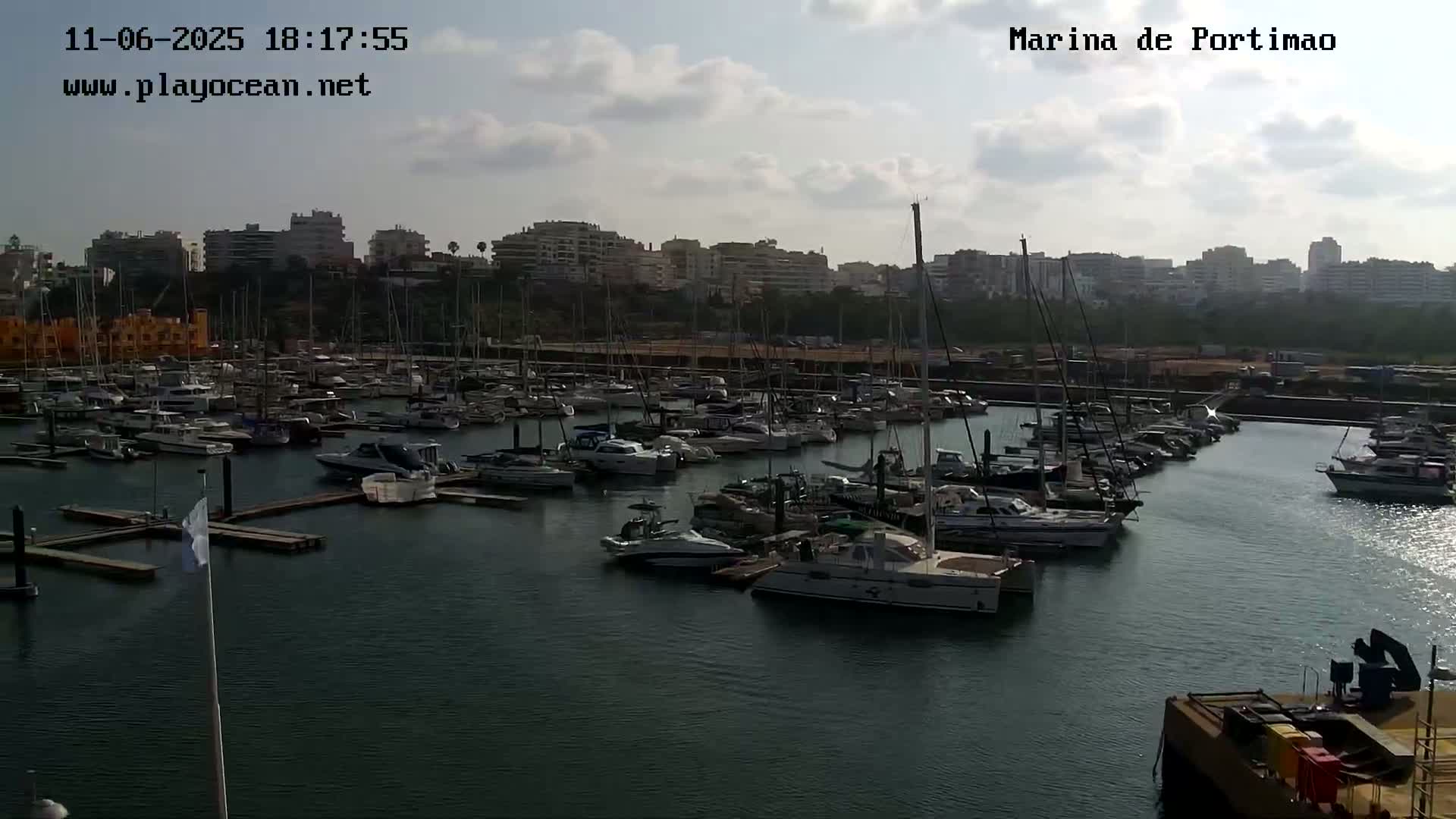 A marina filled with numerous boats and yachts is situated in front of a city skyline under a partly cloudy sky.