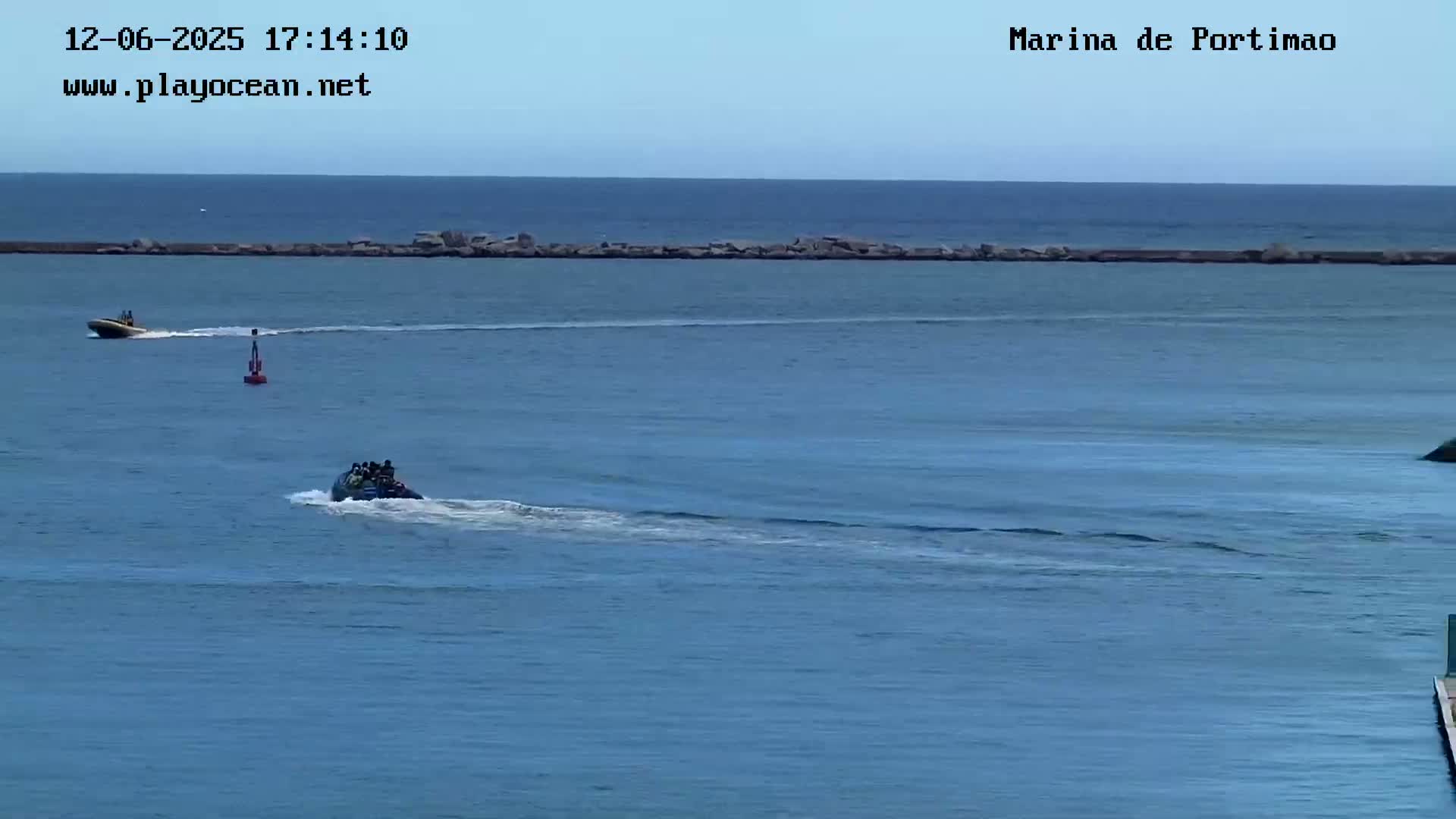 Two jet skis are traveling across a calm blue ocean near a low rock wall under a clear sky.