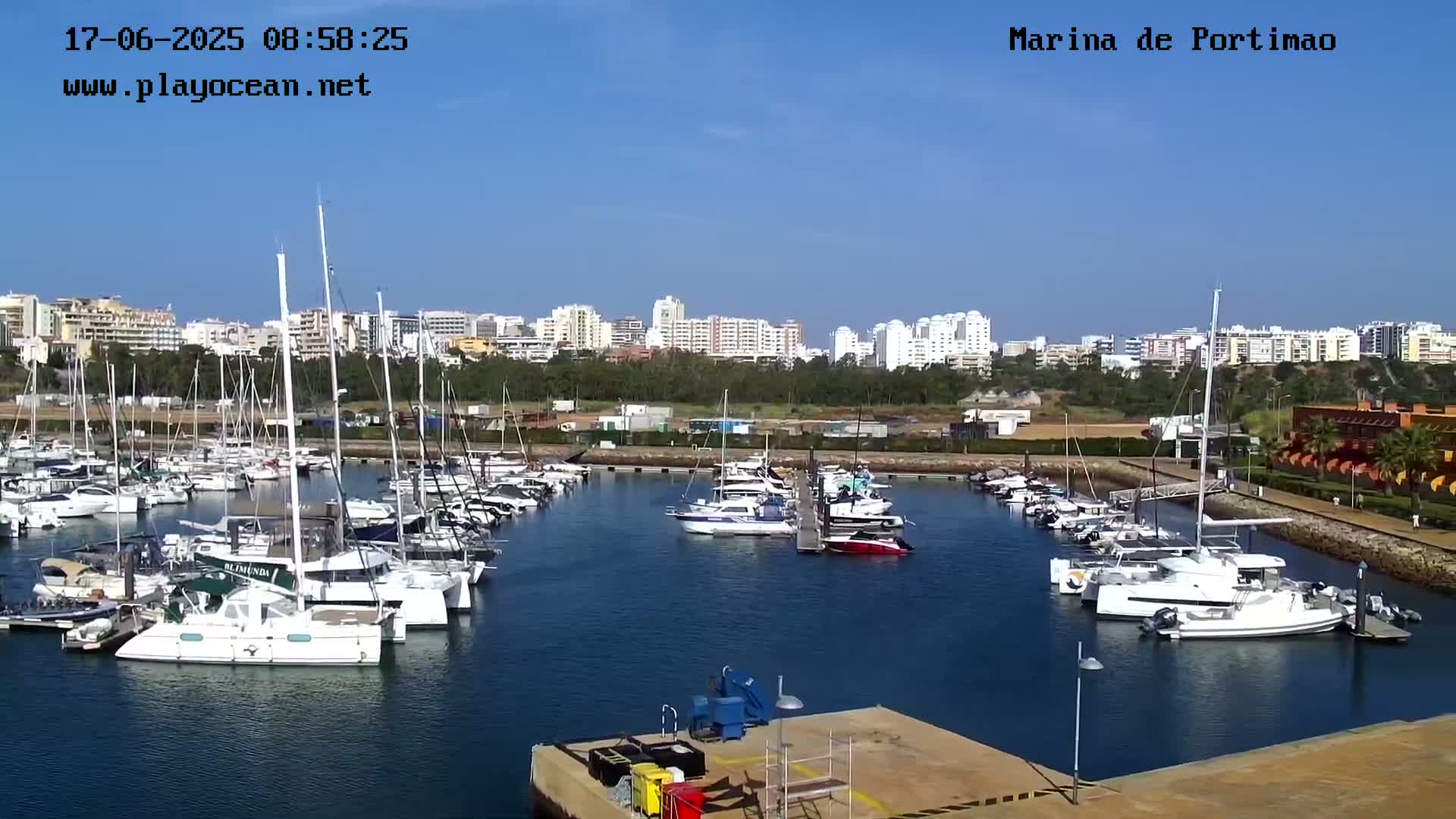A marina filled with numerous sailboats and motorboats is situated on calm water under a clear blue sky, with a cityscape in the background.