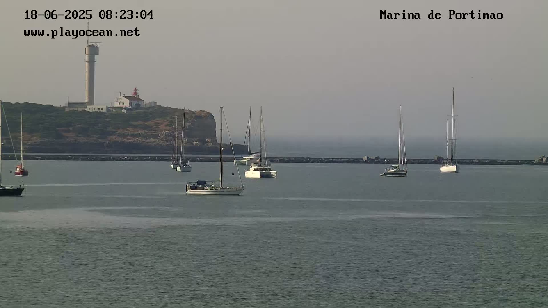 Several sailboats are moored in a calm harbor, with a lighthouse and a tall tower visible on a distant headland under hazy conditions.