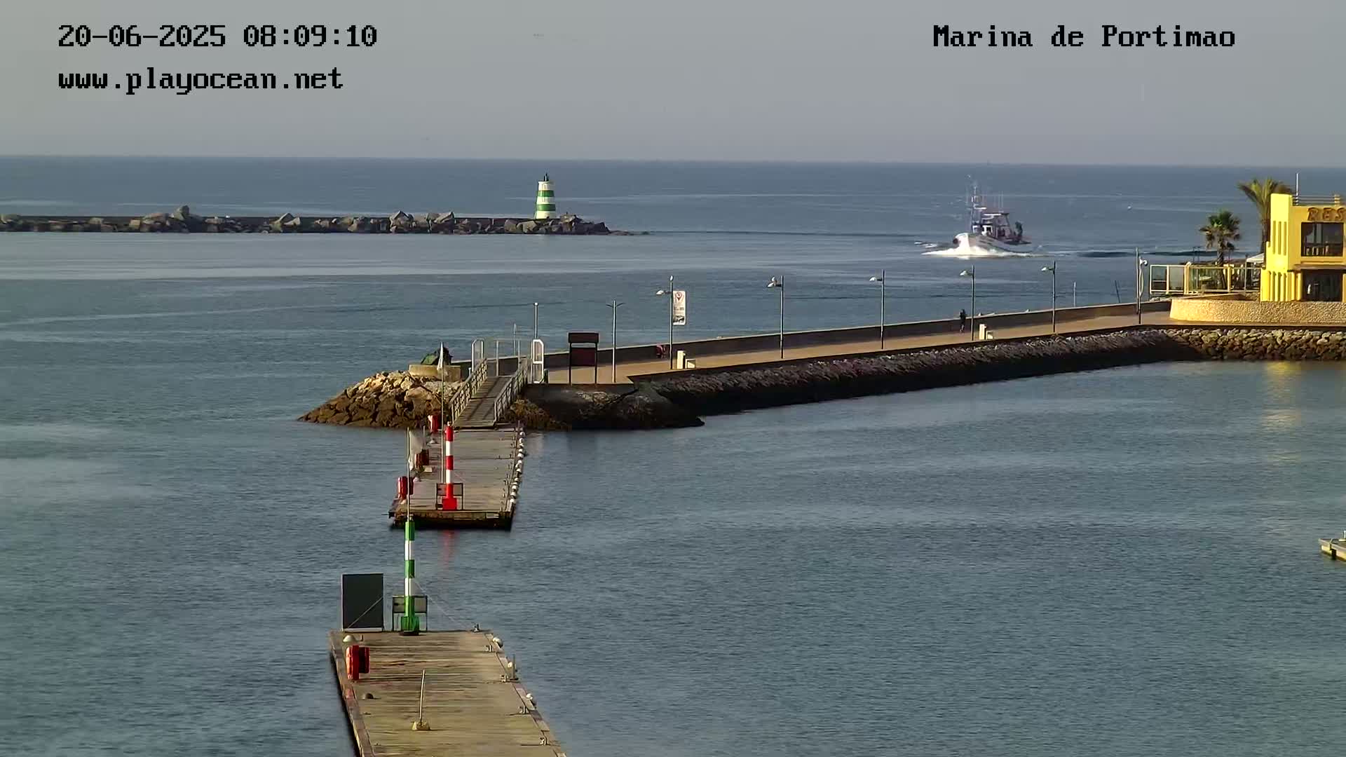 A calm body of water shows a small boat passing by a marina with a walkway and a lighthouse in the distance.