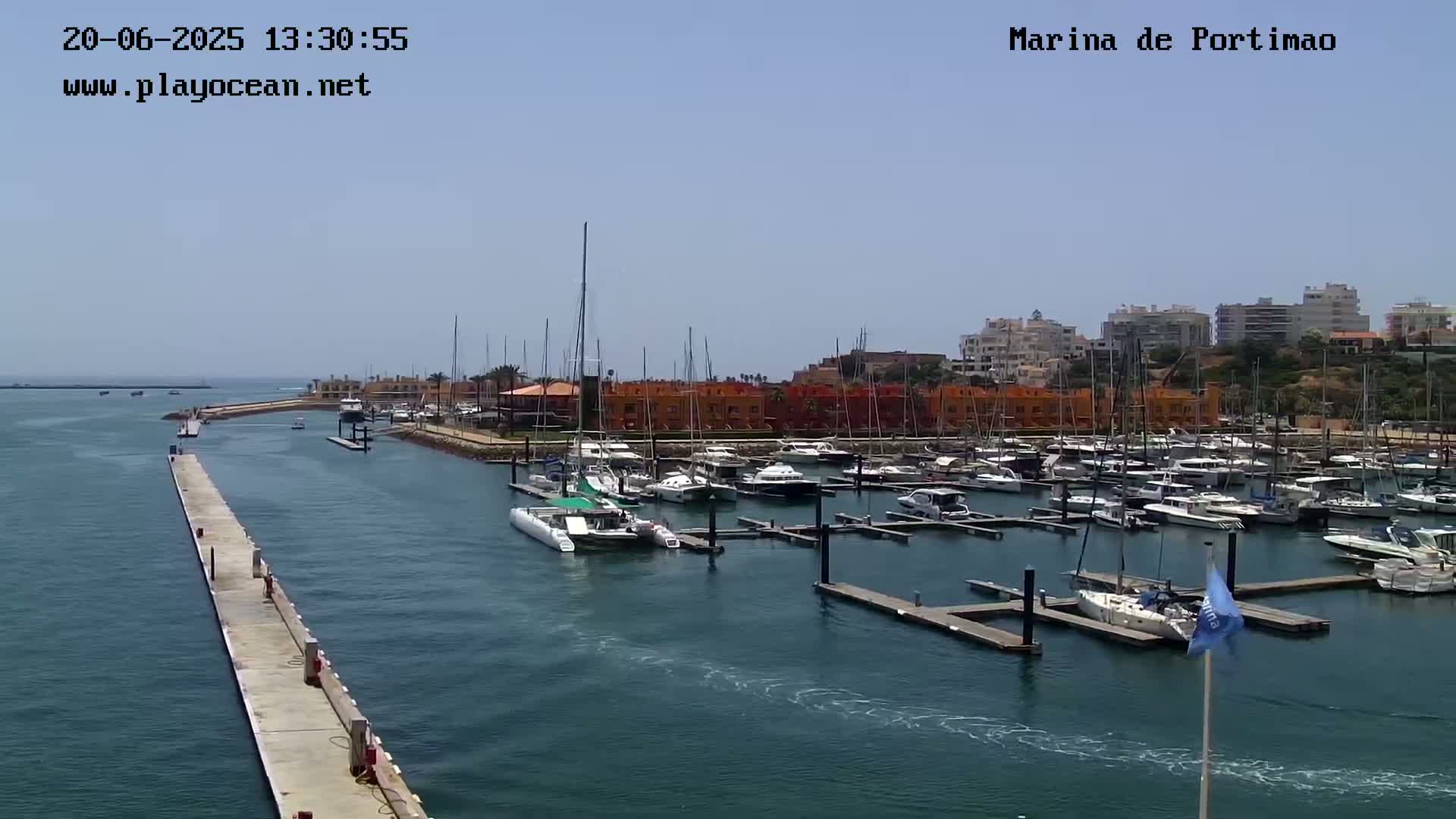 A marina filled with numerous boats and yachts is seen on a sunny day, with a long pier extending into the calm water.