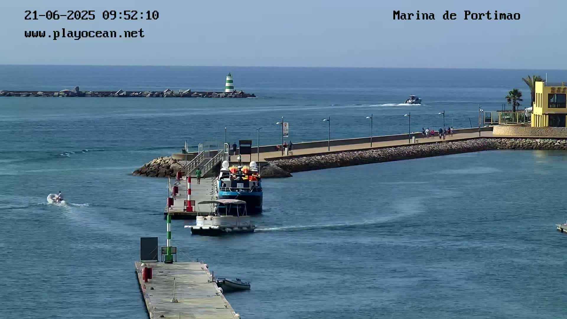 A sunny day shows a marina with two boats at a dock, a person on a jet ski, and people walking along a paved walkway.