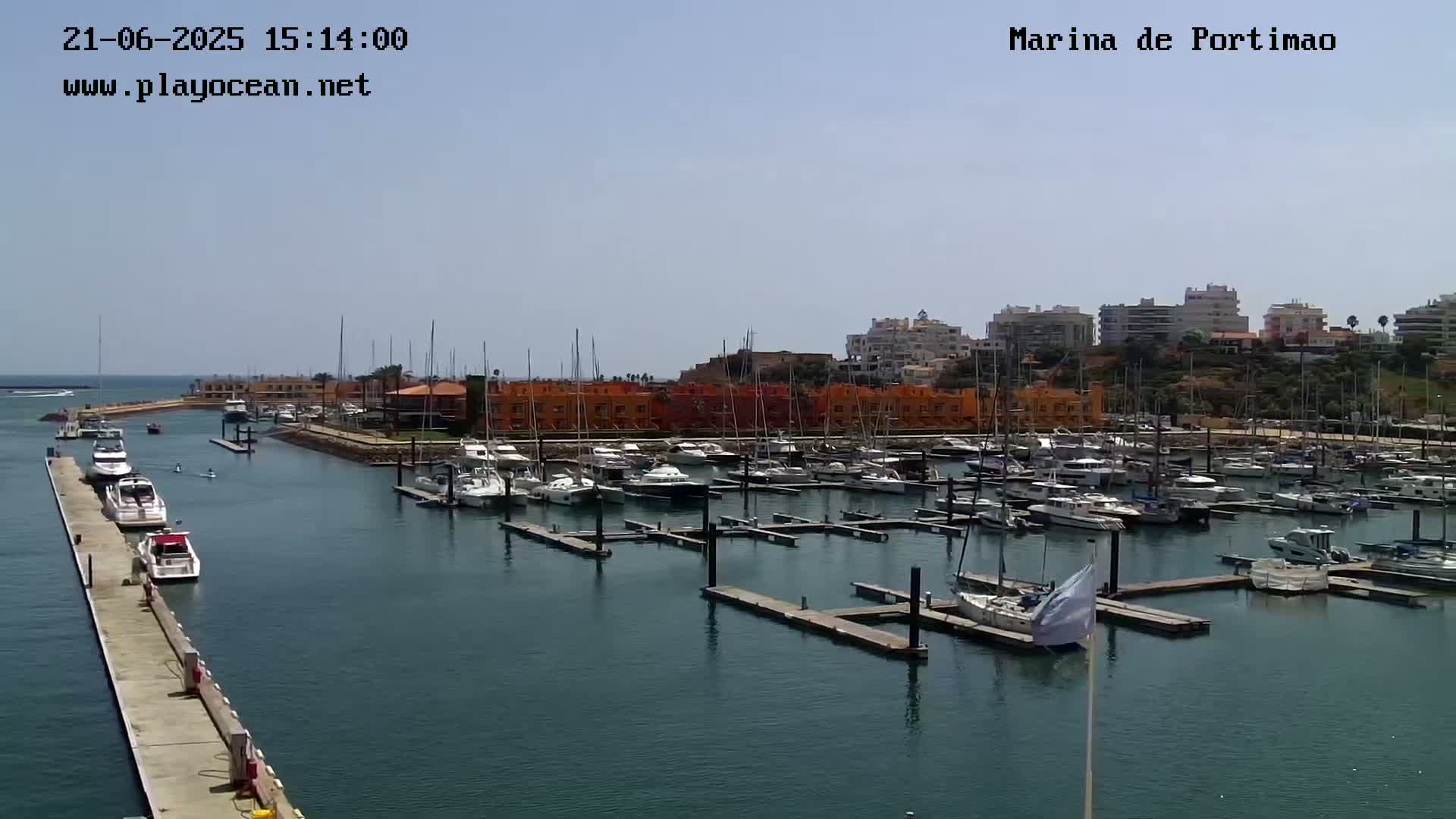 A marina filled with numerous boats is situated on calm, dark-blue water under a clear, sunny sky.