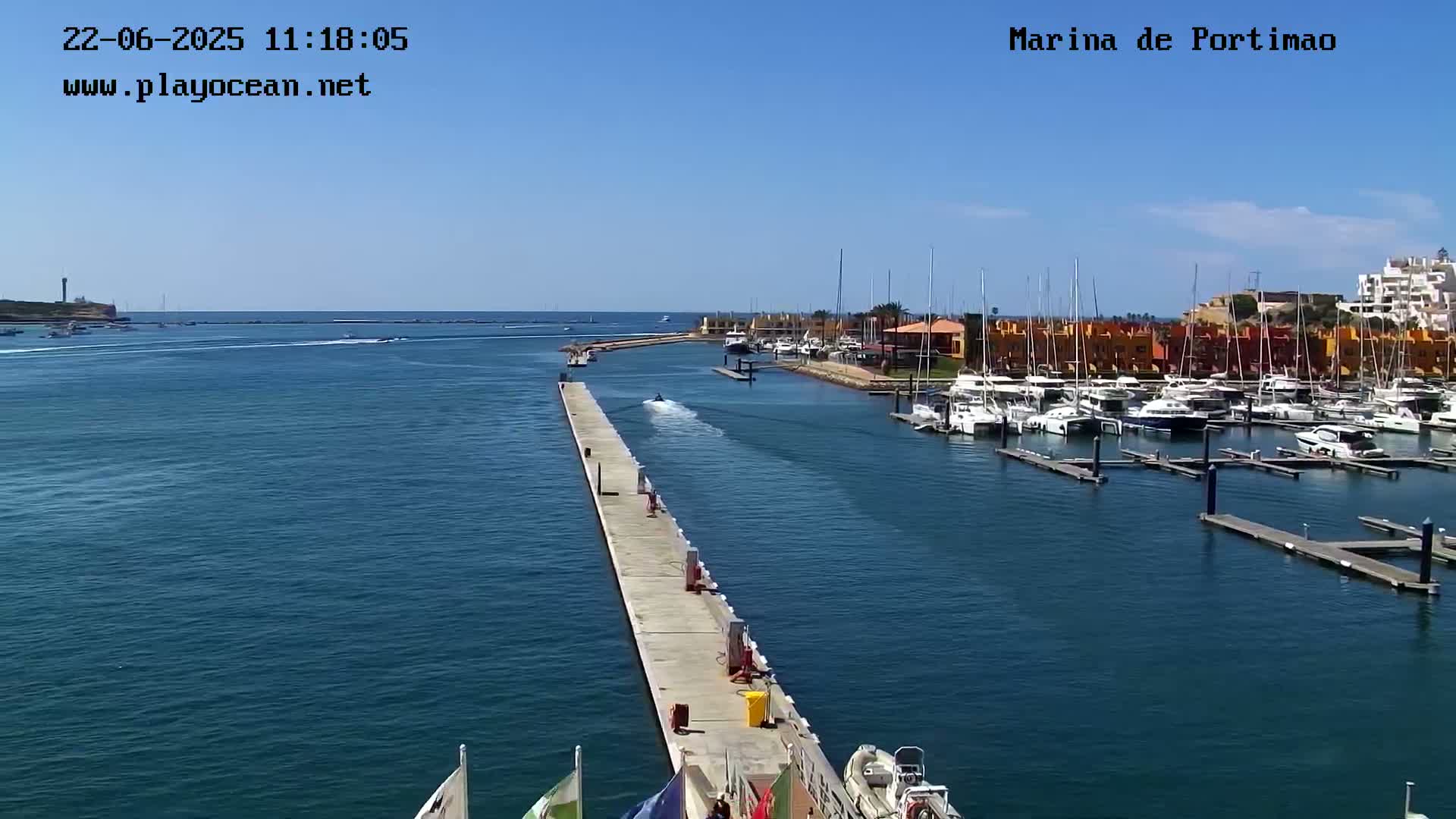 A marina filled with numerous boats and yachts is shown on a sunny day, with a long concrete pier extending into the calm water.