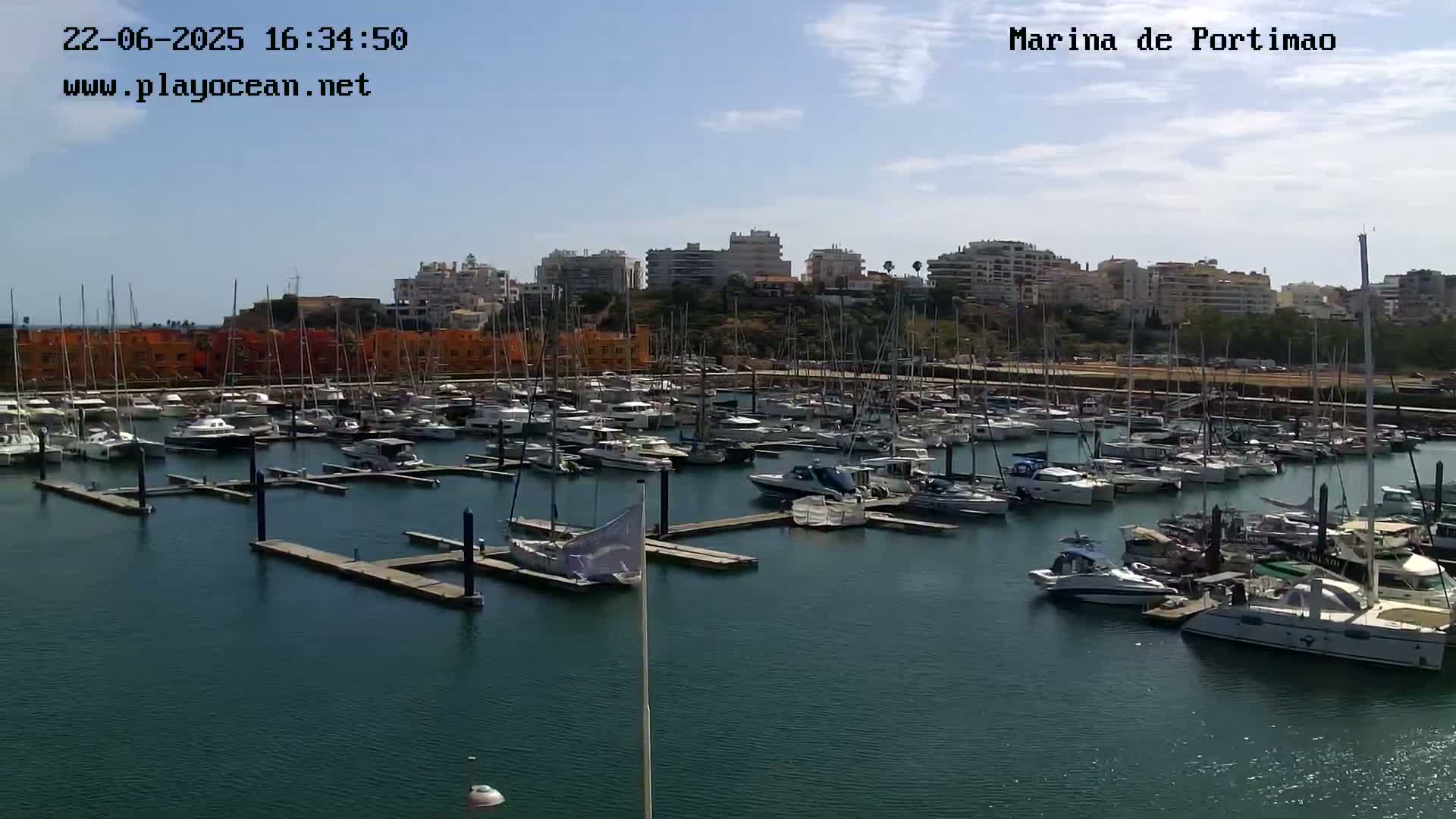 A marina filled with numerous sailboats and motorboats is situated in front of a coastal town under a partly sunny sky.