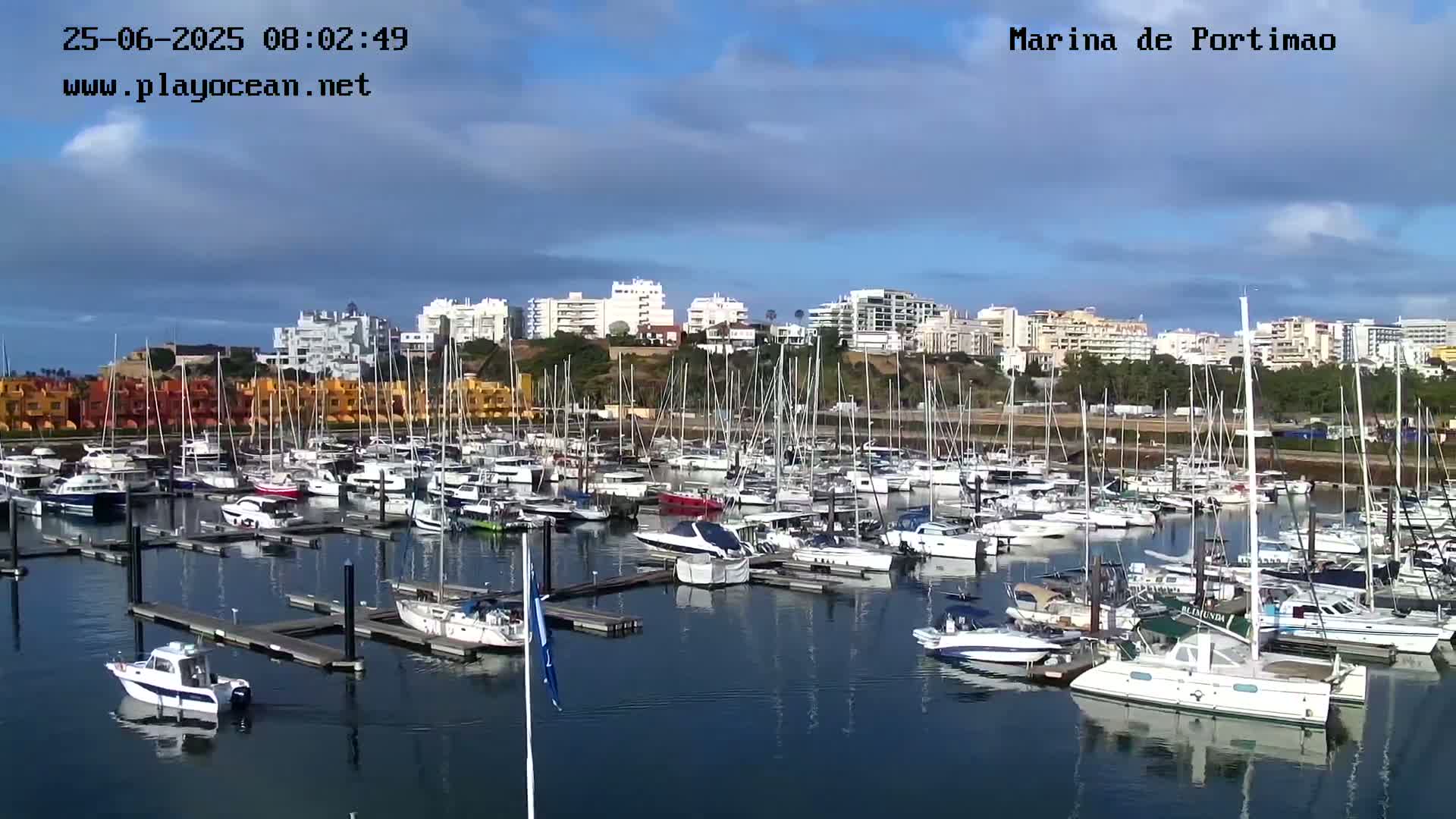A marina filled with numerous boats and yachts is situated under a partly cloudy sky.