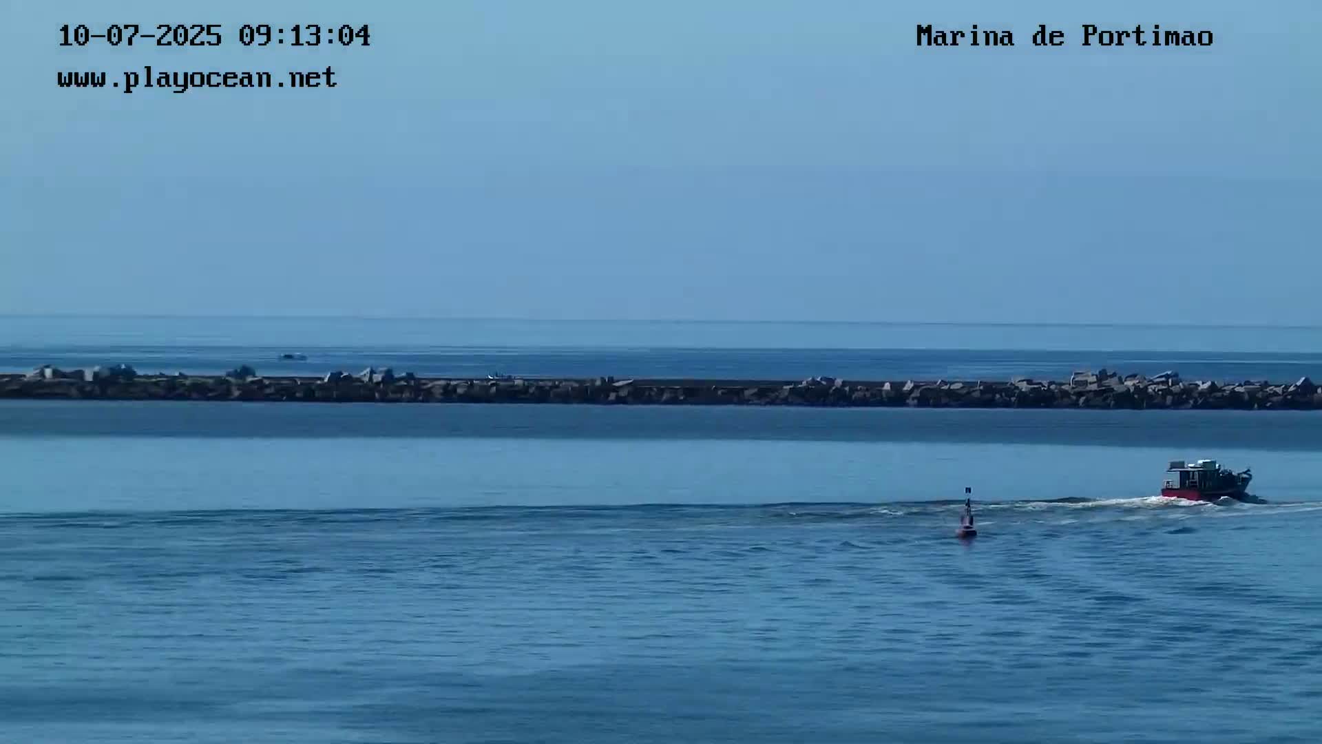 A small boat travels across calm, blue water past a rock jetty under a clear sky.